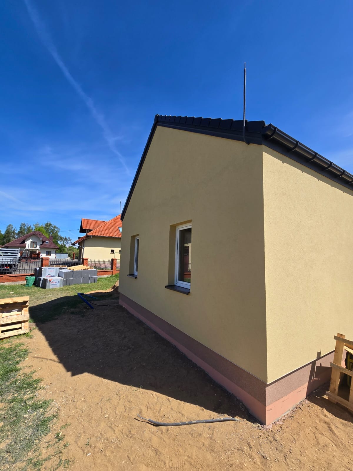 Żółta elewacja domu jednorodzinnego z widocznym ociepleniem fundamentu, dwa okna. Na pierwszym planie piasek i cień. Błękitne niebo.