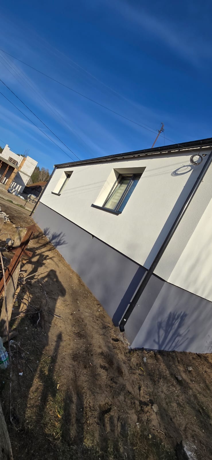 Elewacja budynku po malowaniu z szarą podmurówką i białymi ścianami w słoneczny dzień. Widoczne dwa okna i fragment dachu. W tle budowa innego budynku.