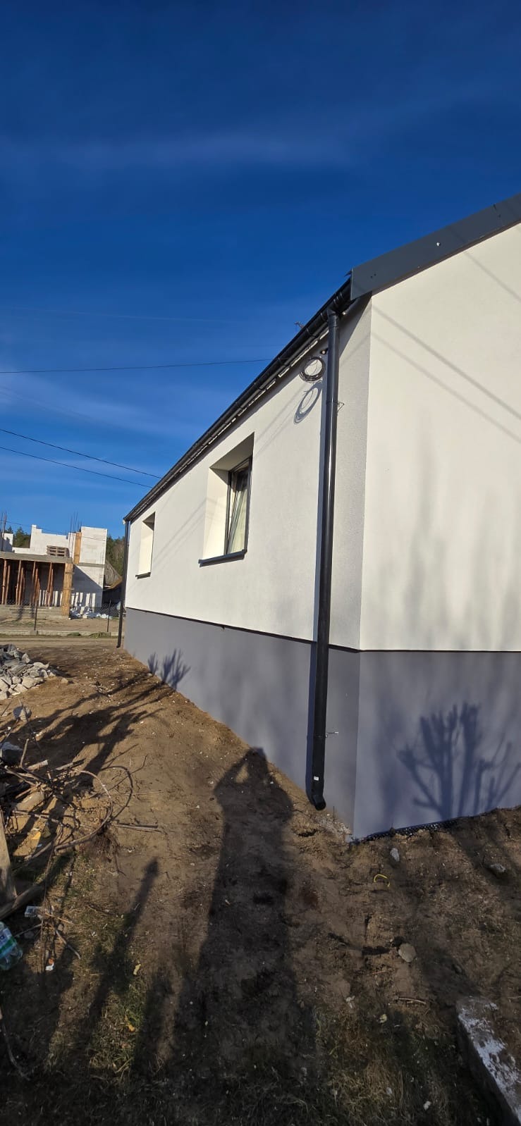 Wykończona elewacja budynku z widocznym systemem rynnowym, dwukolorową fasadą (biało-szara) i oknami. W tle budynek w trakcie budowy.