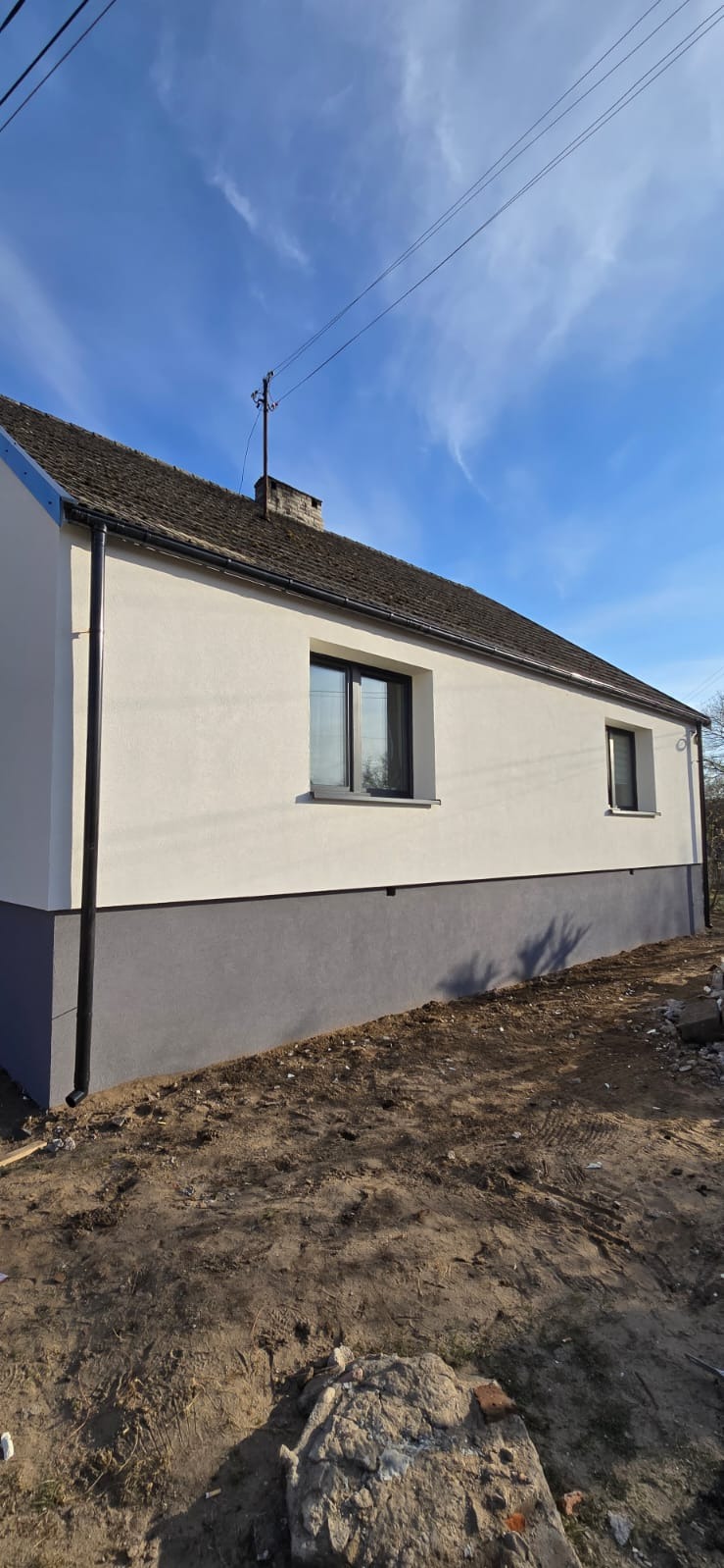 Dwukolorowa elewacja domu jednorodzinnego z ciemnymi oknami, widok z dołu w słoneczny dzień. Elewacja świeżo po malowaniu, z widocznym podziałem na dwie strefy kolorystyczne.