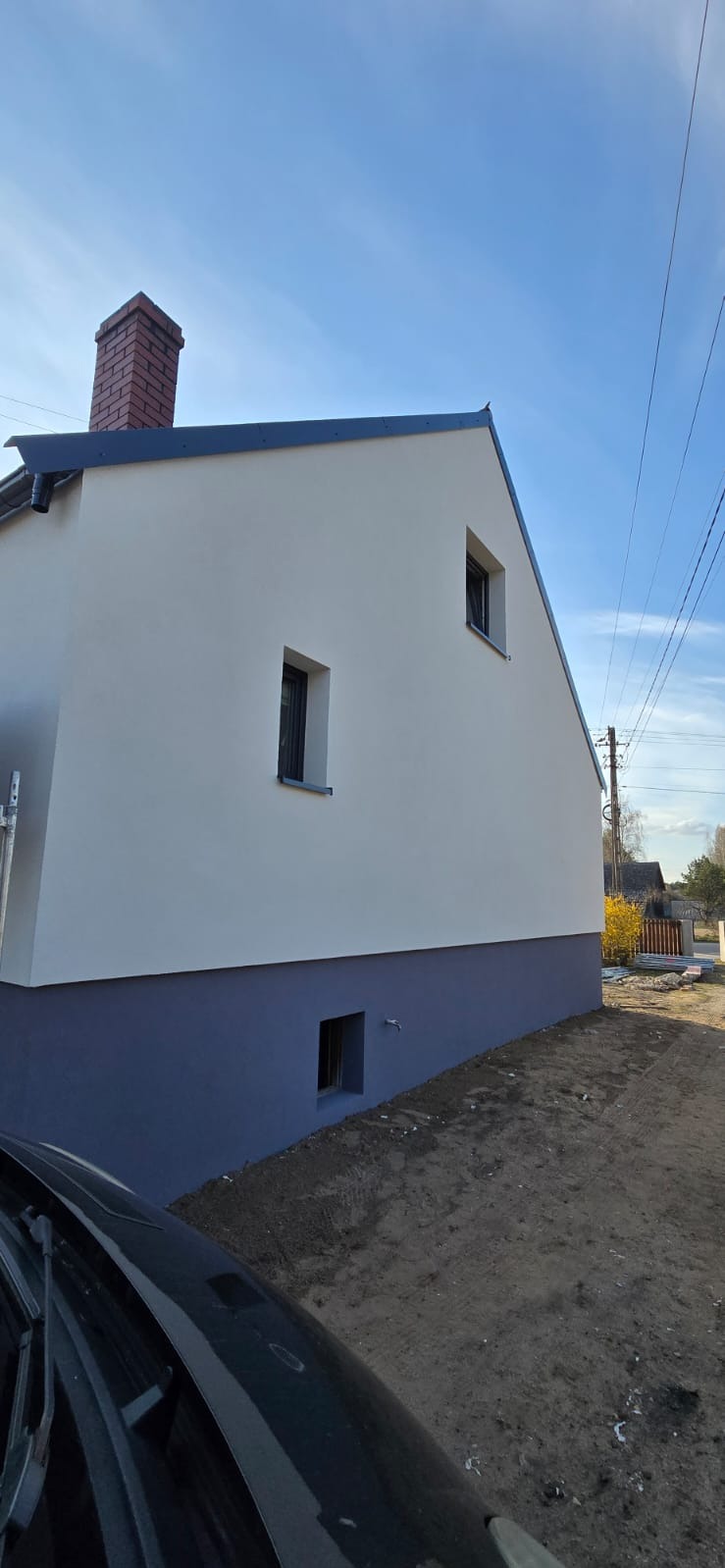 Świeżo pomalowana elewacja domu z ceglanym kominem w słoneczny dzień. Dolna część elewacji w kolorze szarym, reszta biała. Widoczne małe okna.