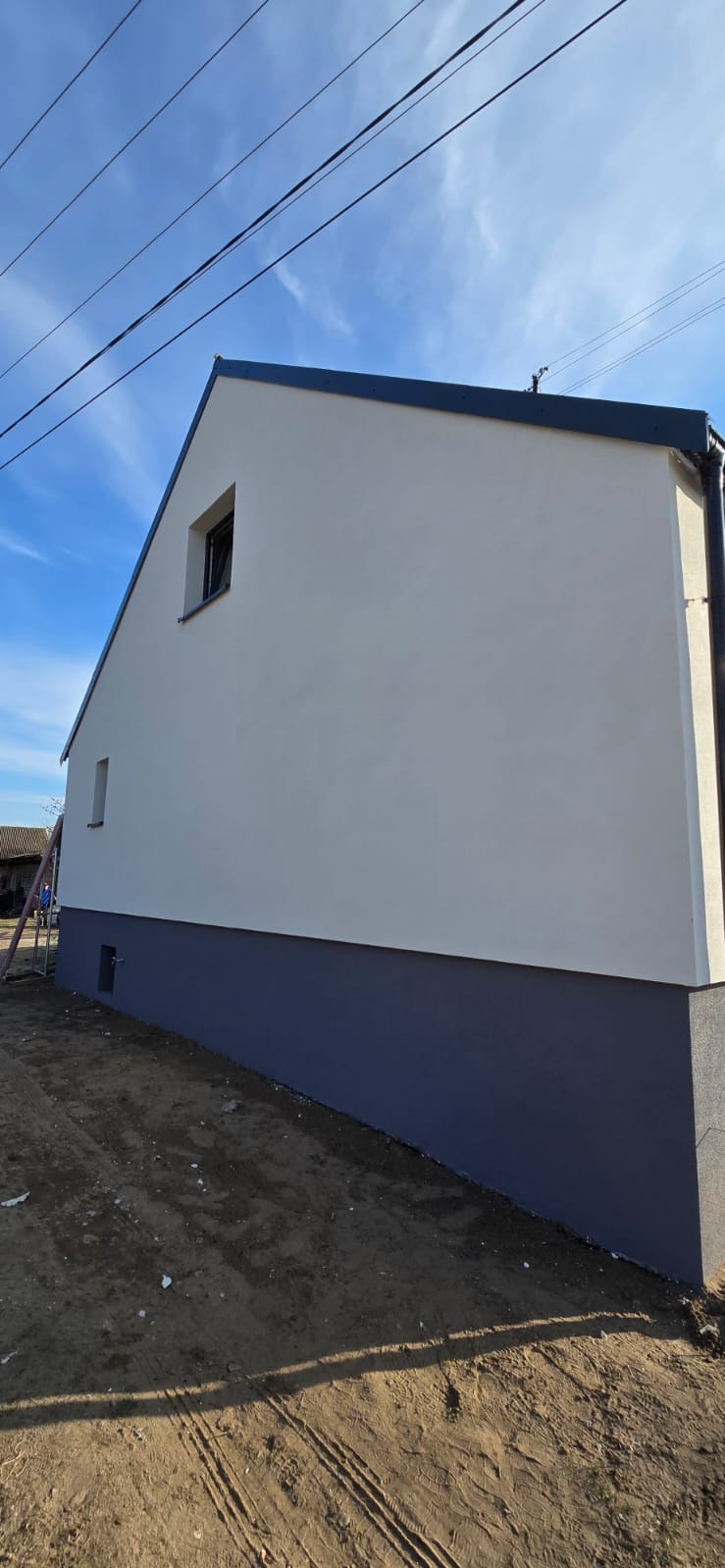 Świeżo pomalowana elewacja domu jednorodzinnego w odcieniach bieli i szarości, widok z boku na tle błękitnego nieba z liniami energetycznymi.