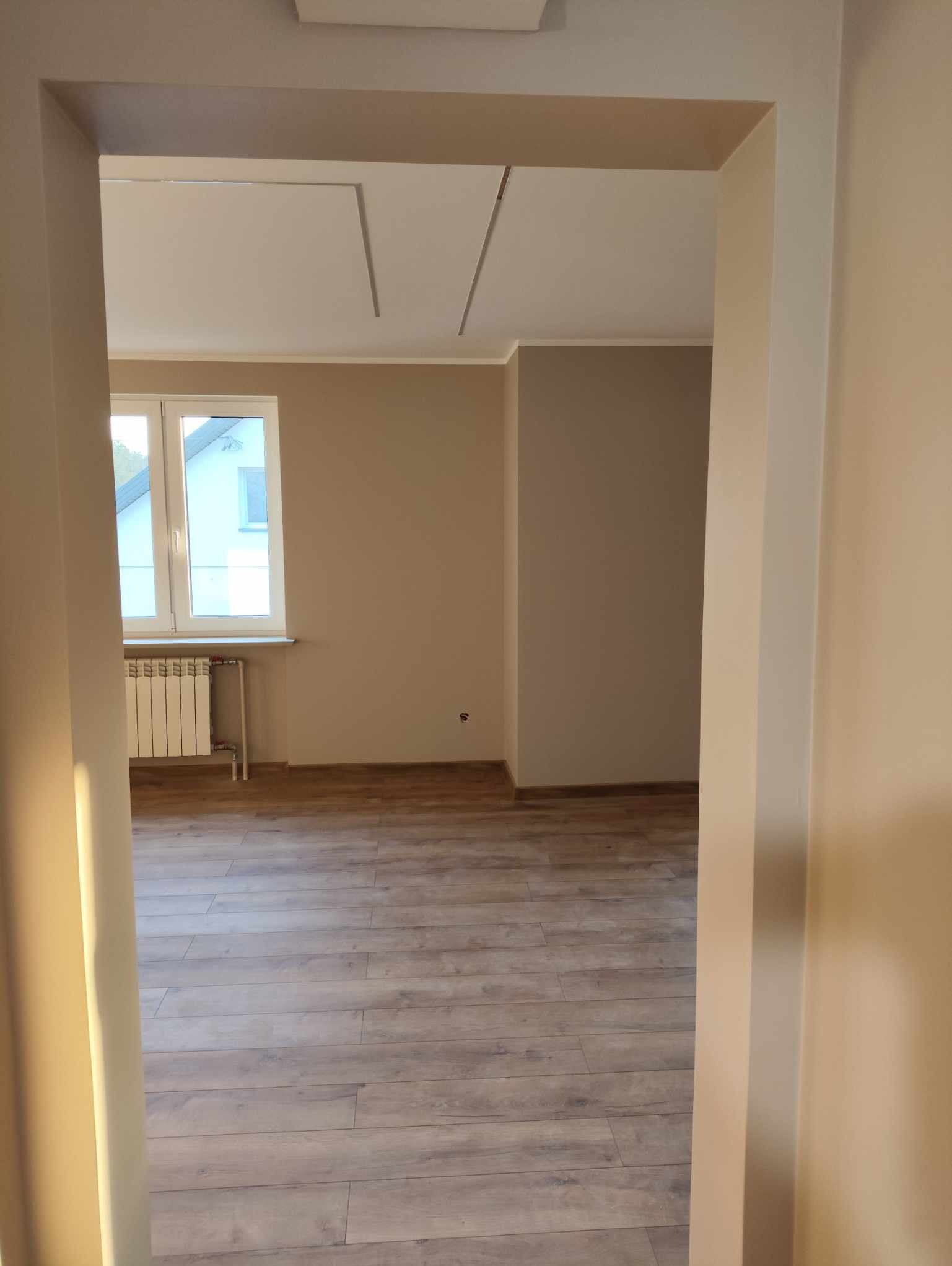 Jasne, świeżo pomalowane wnętrze z drewnianą podłogą, widoczne przez framugę drzwi. Grzejnik pod oknem. Neutralna kolorystyka ścian.
