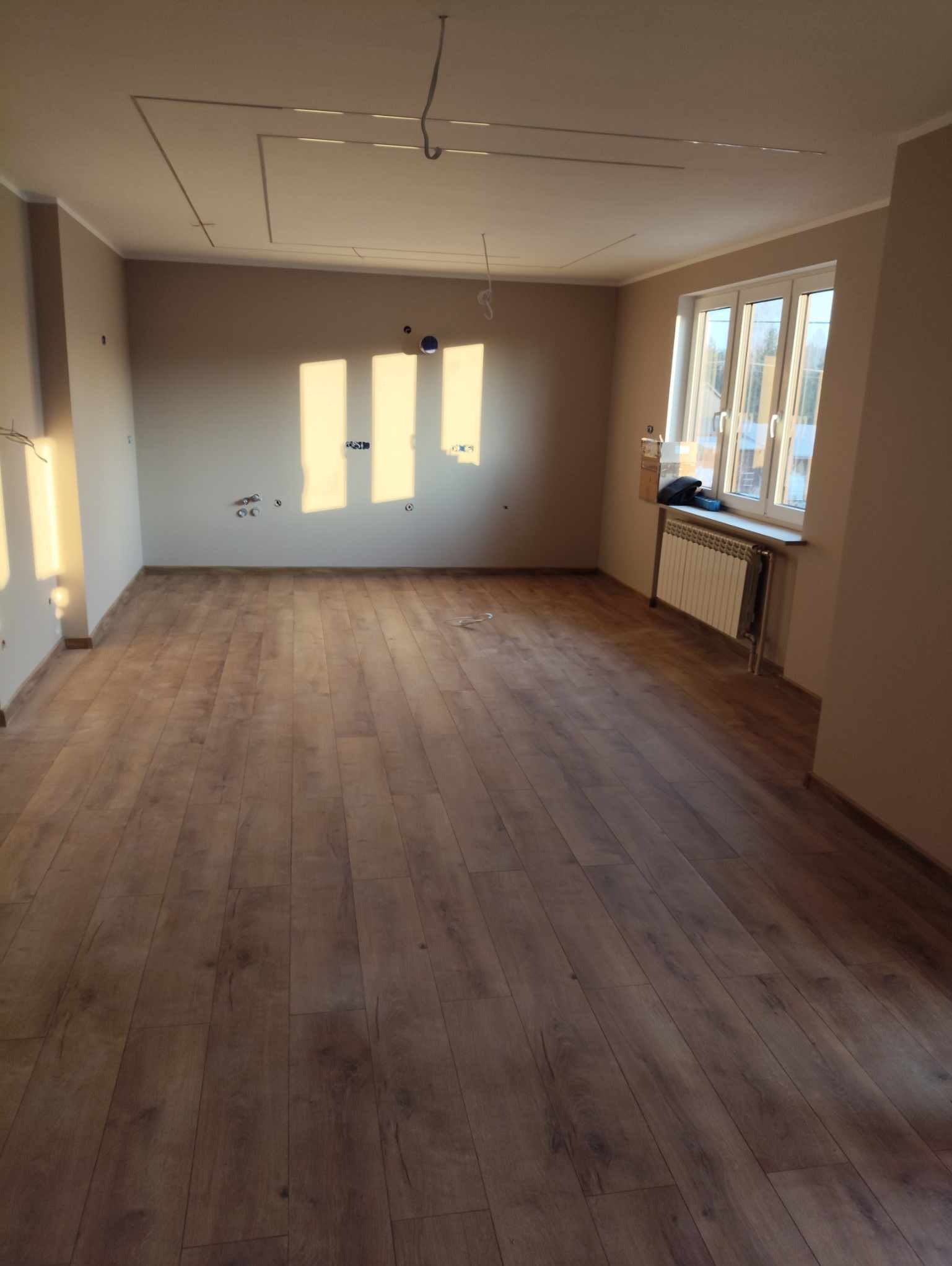 Jasne, przestronne wnętrze po remoncie: drewniana podłoga, świeżo pomalowane ściany, okno z grzejnikiem. Widoczne przygotowania do montażu oświetlenia sufitowego.