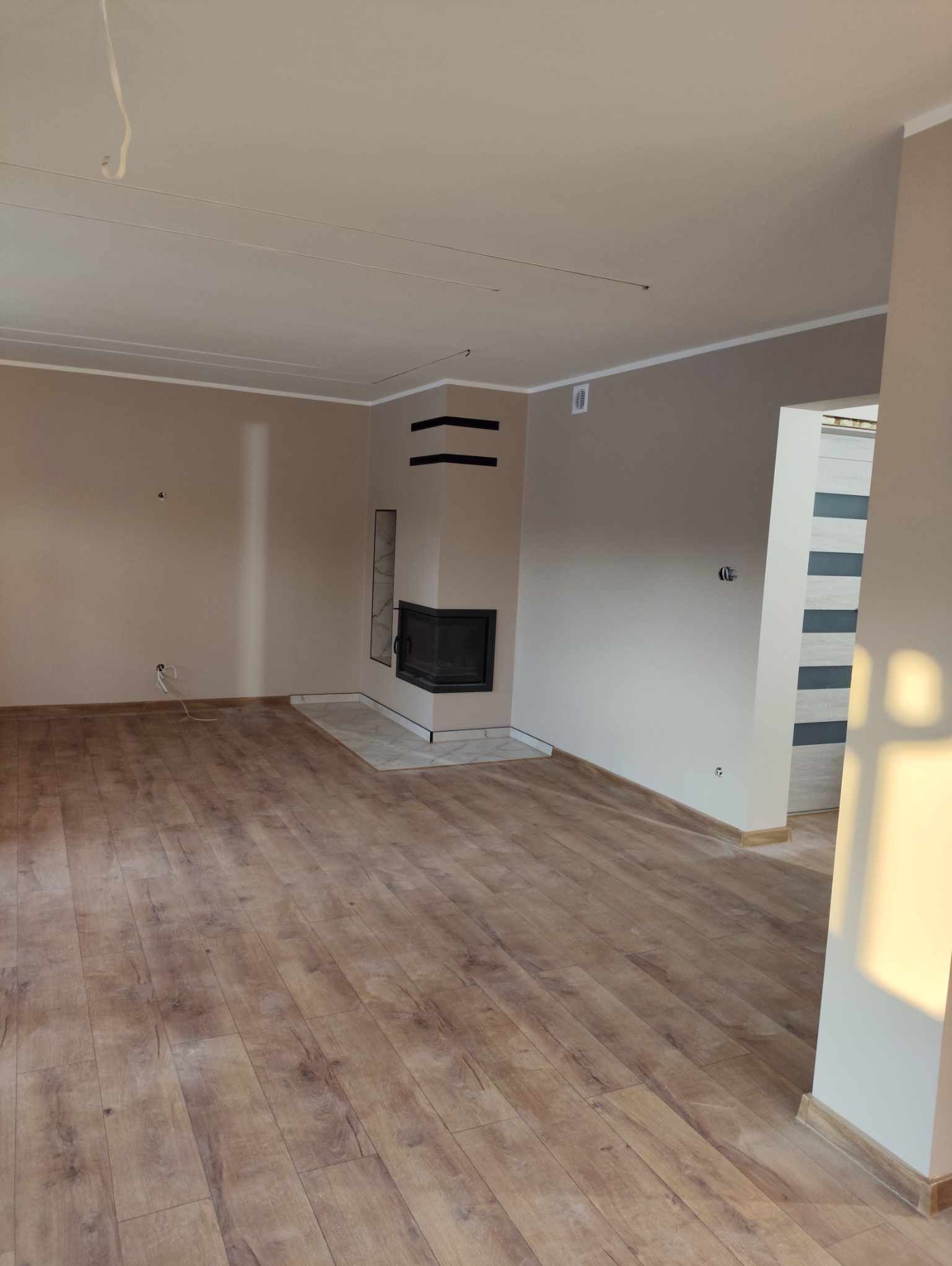 Jasne, przestronne wnętrze z kominkiem i drewnianą podłogą. Ściany w odcieniach beżu, minimalistyczny design. Widoczne gniazdka elektryczne i listwy przypodłogowe.