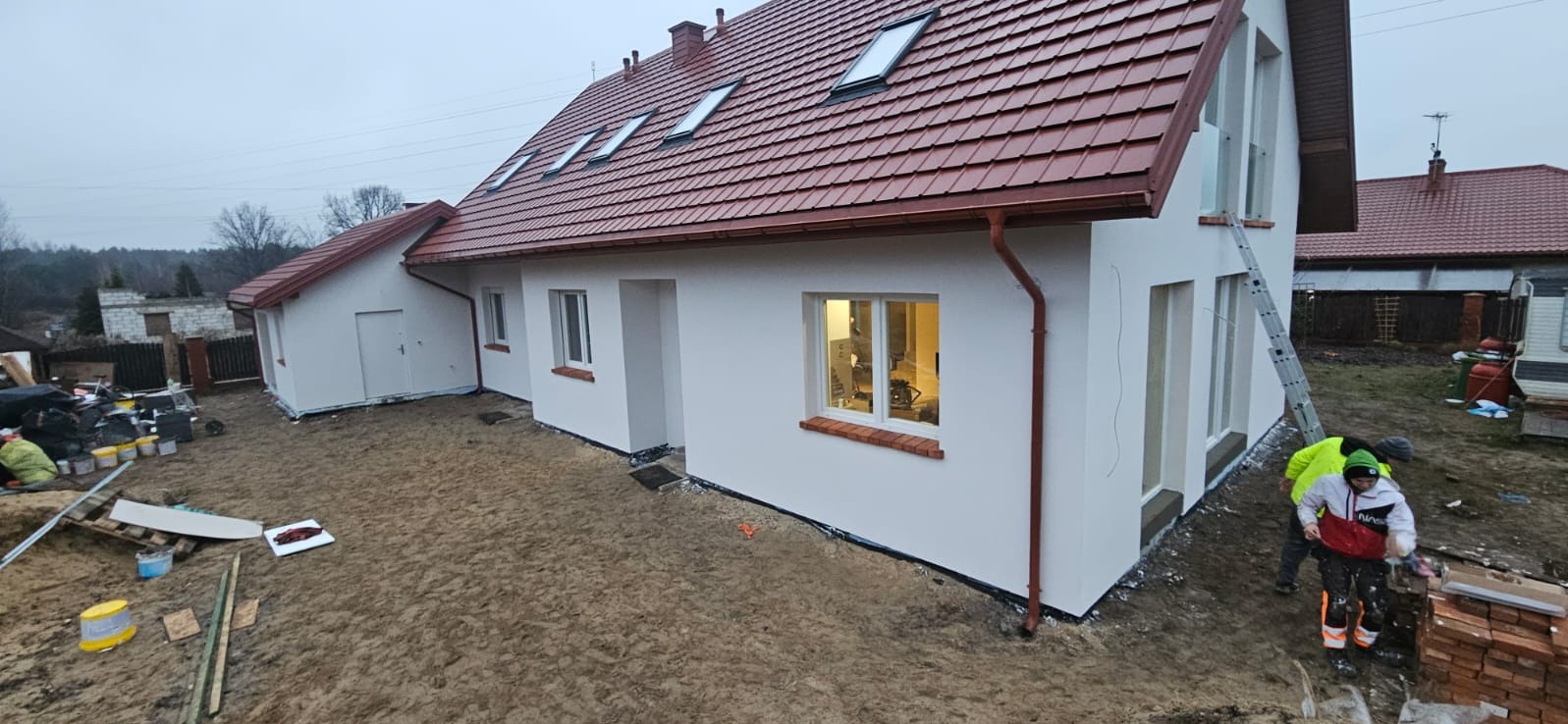Biała elewacja domu z brązowym dachem i oknami, drabina oparta o ścianę, widoczni pracownicy w trakcie prac wykończeniowych, teren wokół domu przygotowany pod zagospodarowanie.