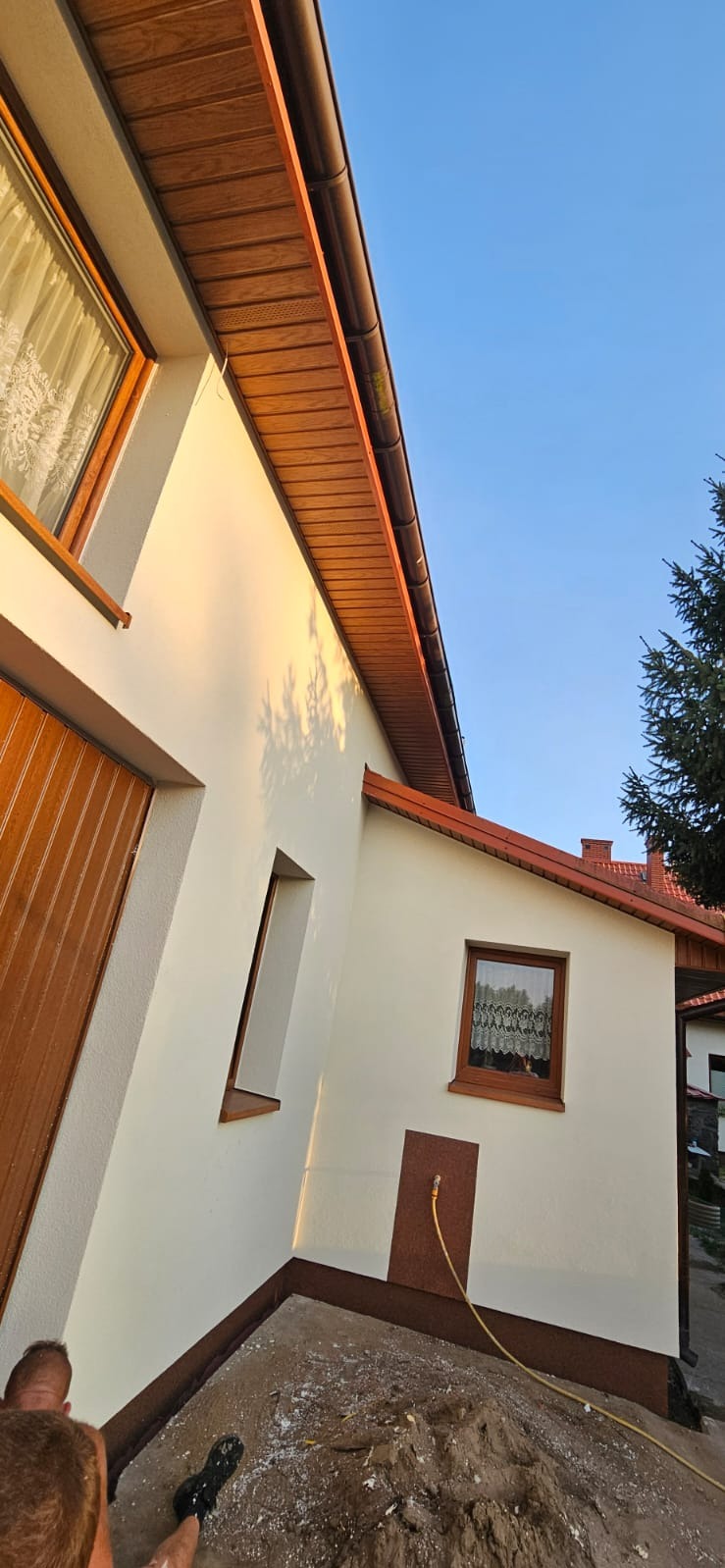 Jasna elewacja domu z drewnianymi elementami wykończeniowymi i widocznym fragmentem dachu. W tle błękitne niebo i zieleń drzew. Widoczny wąż ogrodowy.