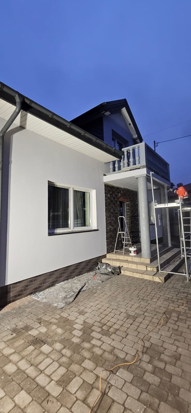 Dom po renowacji elewacji z szarą fasadą, widoczne rusztowania i drabina. Część elewacji wykończona kamieniem dekoracyjnym, kostka brukowa na podjeździe.