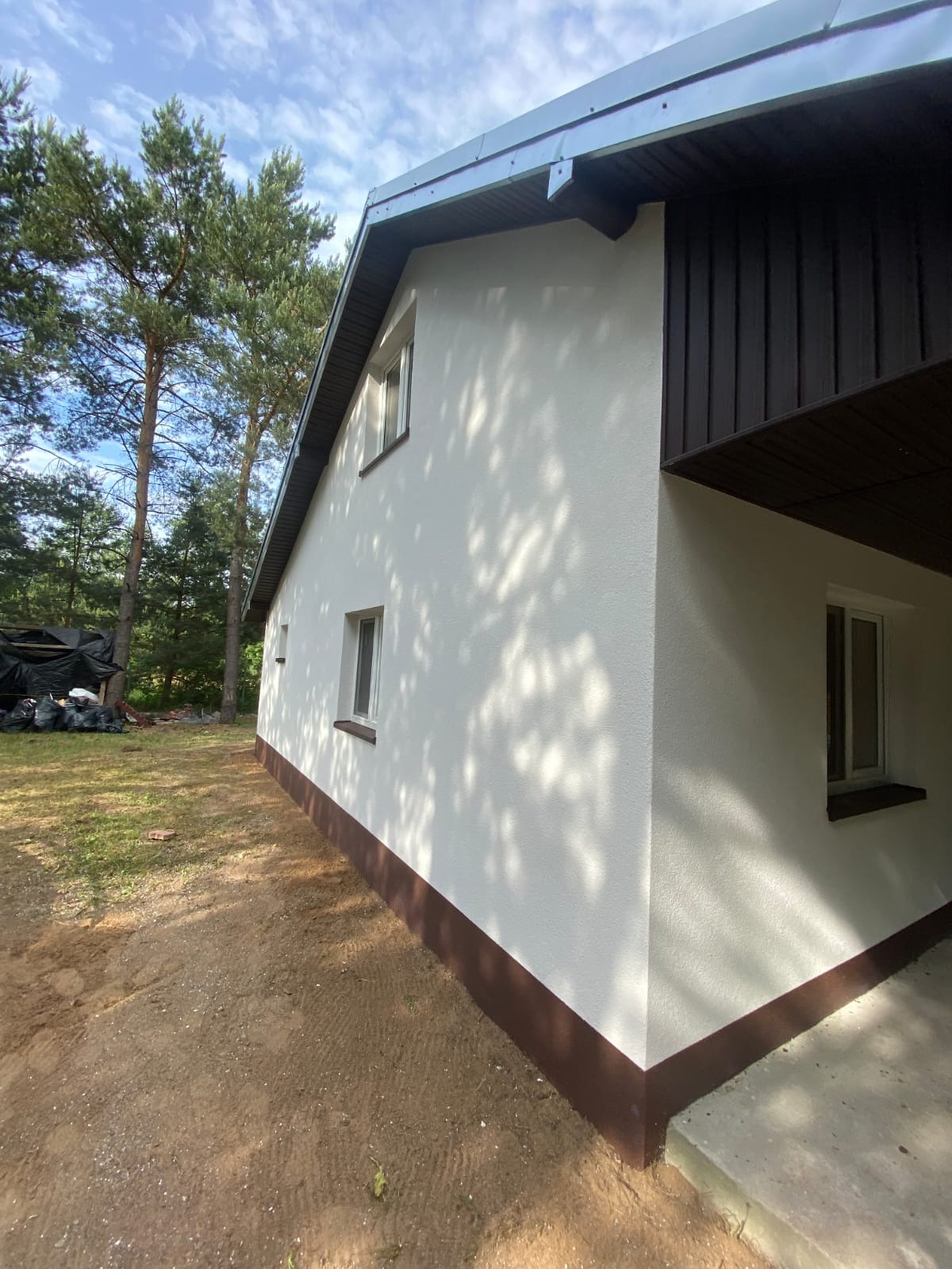 Wykończona elewacja domu jednorodzinnego w Ostrówku. Kremowa ściana z brązowymi akcentami, widok z boku, drzewa w tle, błękitne niebo.