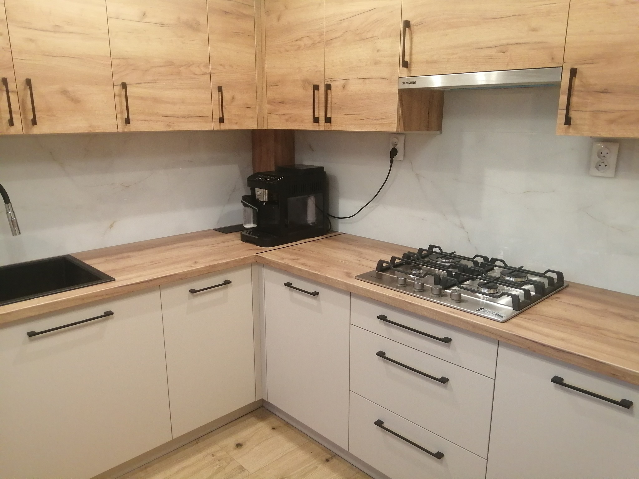 Jasne, drewnopodobne szafki kuchenne z czarnymi uchwytami, marmurowy backsplash, czarny zlew i płyta gazowa. Widoczny ekspres do kawy i gniazdko elektryczne.