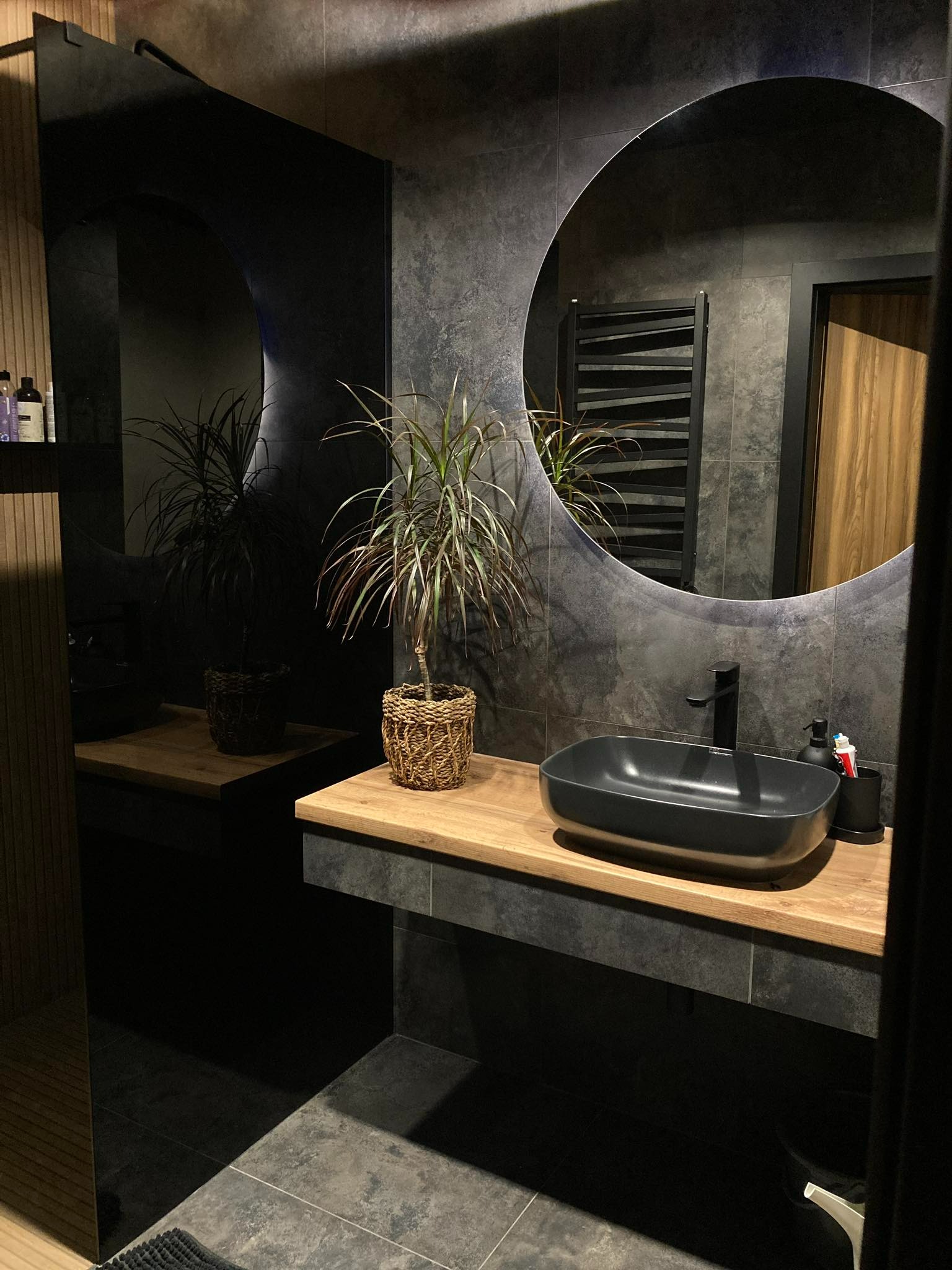 Nowoczesna łazienka z okrągłym lustrem z podświetleniem LED, czarną umywalką i drewnianym blatem. Minimalistyczny design i eleganckie wykończenie.
