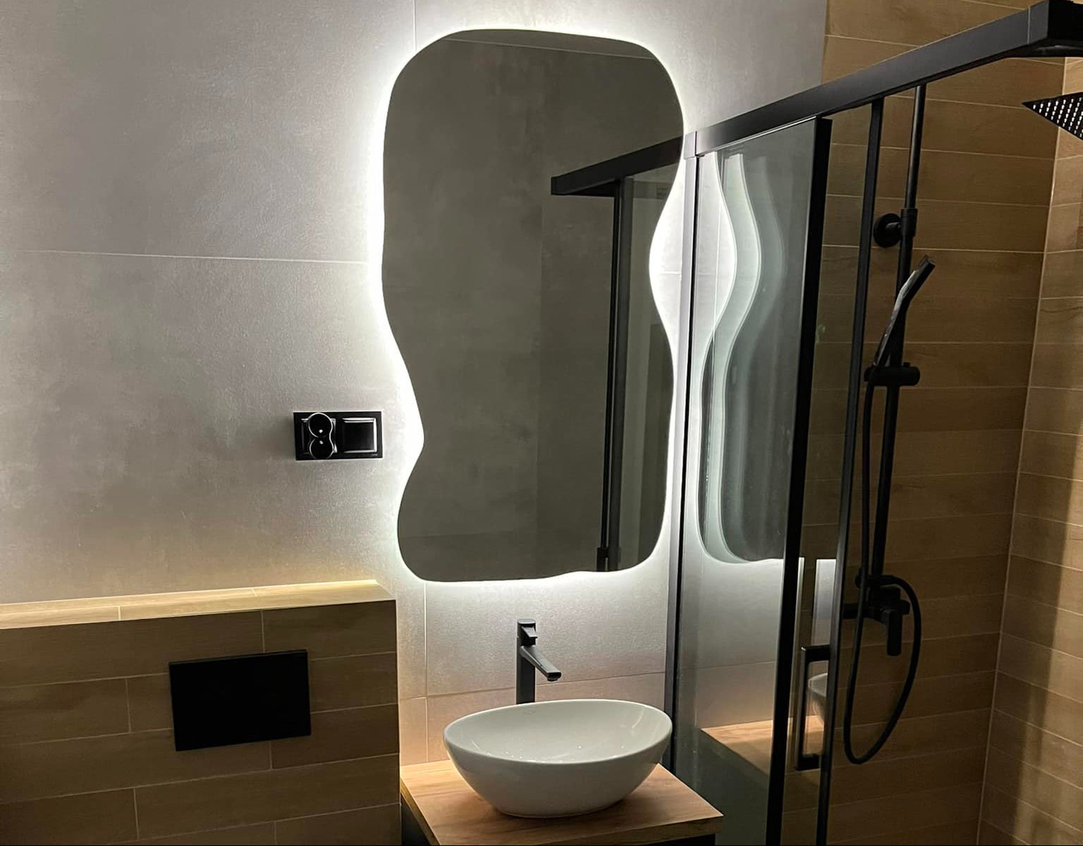 Nowoczesna łazienka z podświetlanym, asymetrycznym lustrem i kabiną prysznicową z ozdobną szybą. Elementy w kolorze czarnym, płytki w odcieniach szarości i brązu.