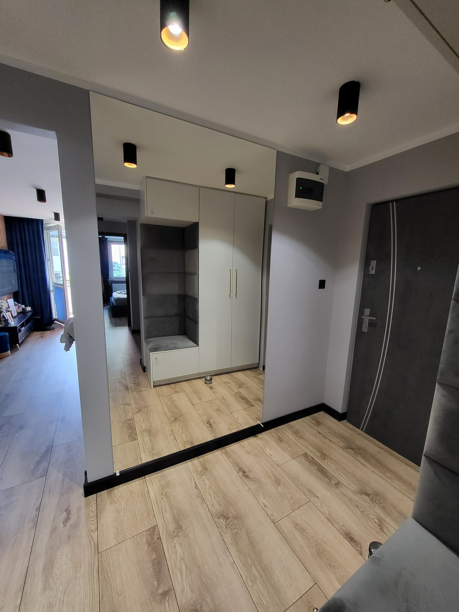 Duże lustro w przedpokoju z wbudowaną szafą i siedziskiem, czarne listwy przypodłogowe, panele podłogowe imitujące drewno, oświetlenie punktowe.