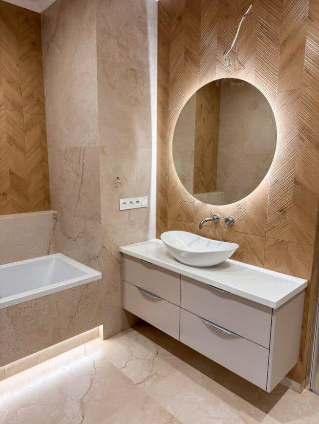 Nowoczesna łazienka z okrągłym lustrem LED, umywalką nablatową i drewnopodobną okładziną ściany w jodełkę. Widoczna wanna i podświetlenie pod szafką.
