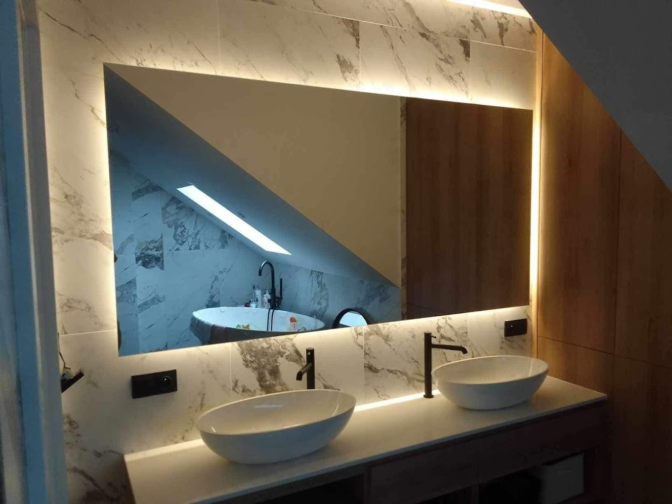 Łazienka z dwoma umywalkami i lustrem z podświetleniem LED, odbijającym skos dachu i okno. Marmurowe płytki na ścianie.