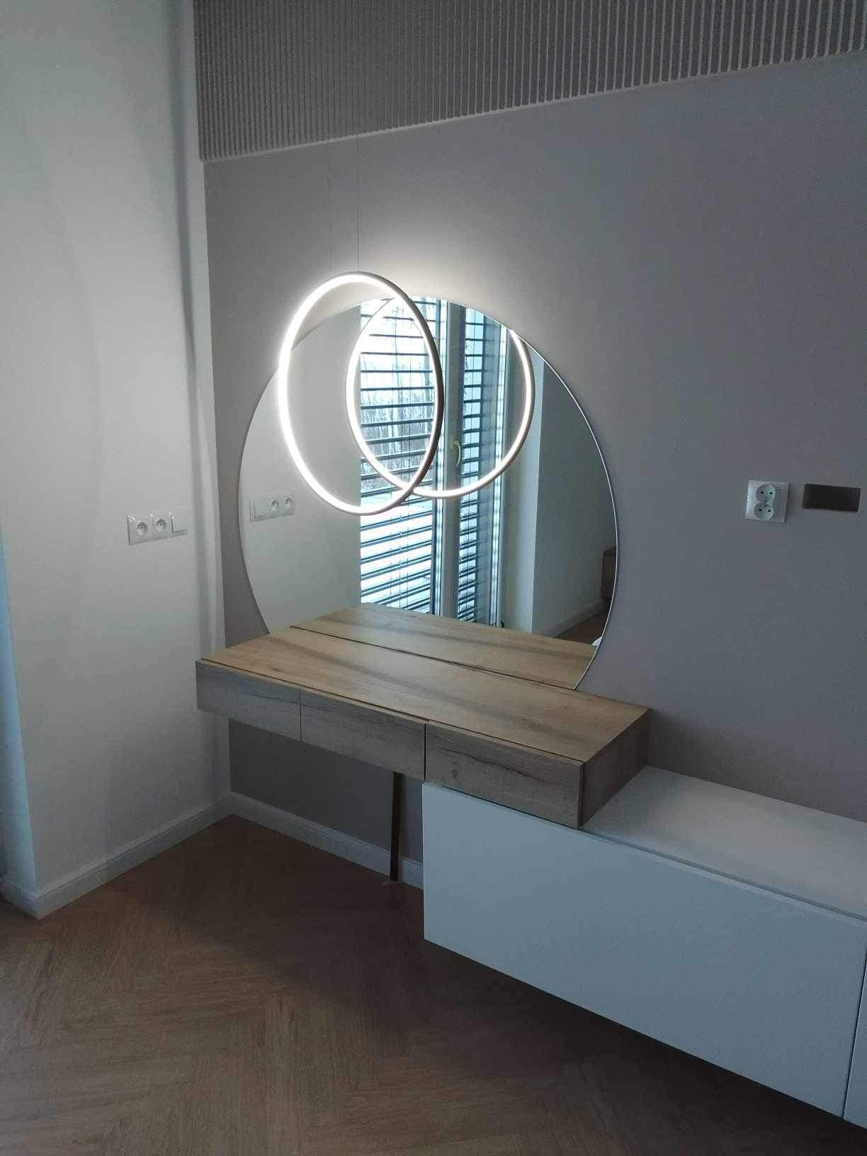 Okrągłe lustro z podświetleniem LED nad drewnianą toaletką w nowoczesnym wnętrzu. Widoczne gniazdka elektryczne na ścianie i parkiet na podłodze.