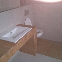 Ernest - Wykończona łazienka z umywalką wpuszczoną w blat z płytek imitujących drewno, toaleta w tle, jasne płytki na ścianach i podłodze, surowy fragment ściany.