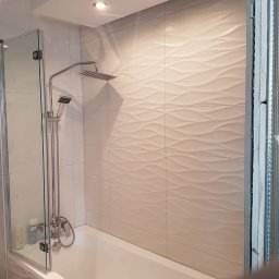 Ernest - Nowoczesna łazienka z białą wanną, szklanym parawanem i deszczownicą. Ściany wyłożone płytkami z delikatnym, falistym wzorem. Oświetlenie punktowe w suficie.