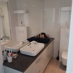 Ernest - Nowoczesna łazienka z dwoma białymi umywalkami na ciemnym blacie, duże lustro, białe płytki i szafki. Widoczna toaleta i akcesoria łazienkowe.