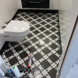 Łazienka po remoncie: czarno-biała mozaika na podłodze, wisząca toaleta i szafka z umywalką. Widoczne narzędzia glazurnicze.