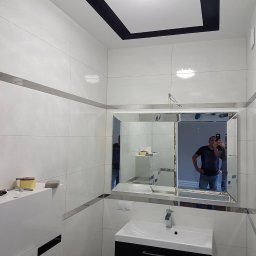 Ernest - Biała łazienka z czarnymi akcentami: kafelki, sufit. Lustro z fazowanymi krawędziami odbija osobę. Wisząca szafka z umywalką w kolorze czarnym.