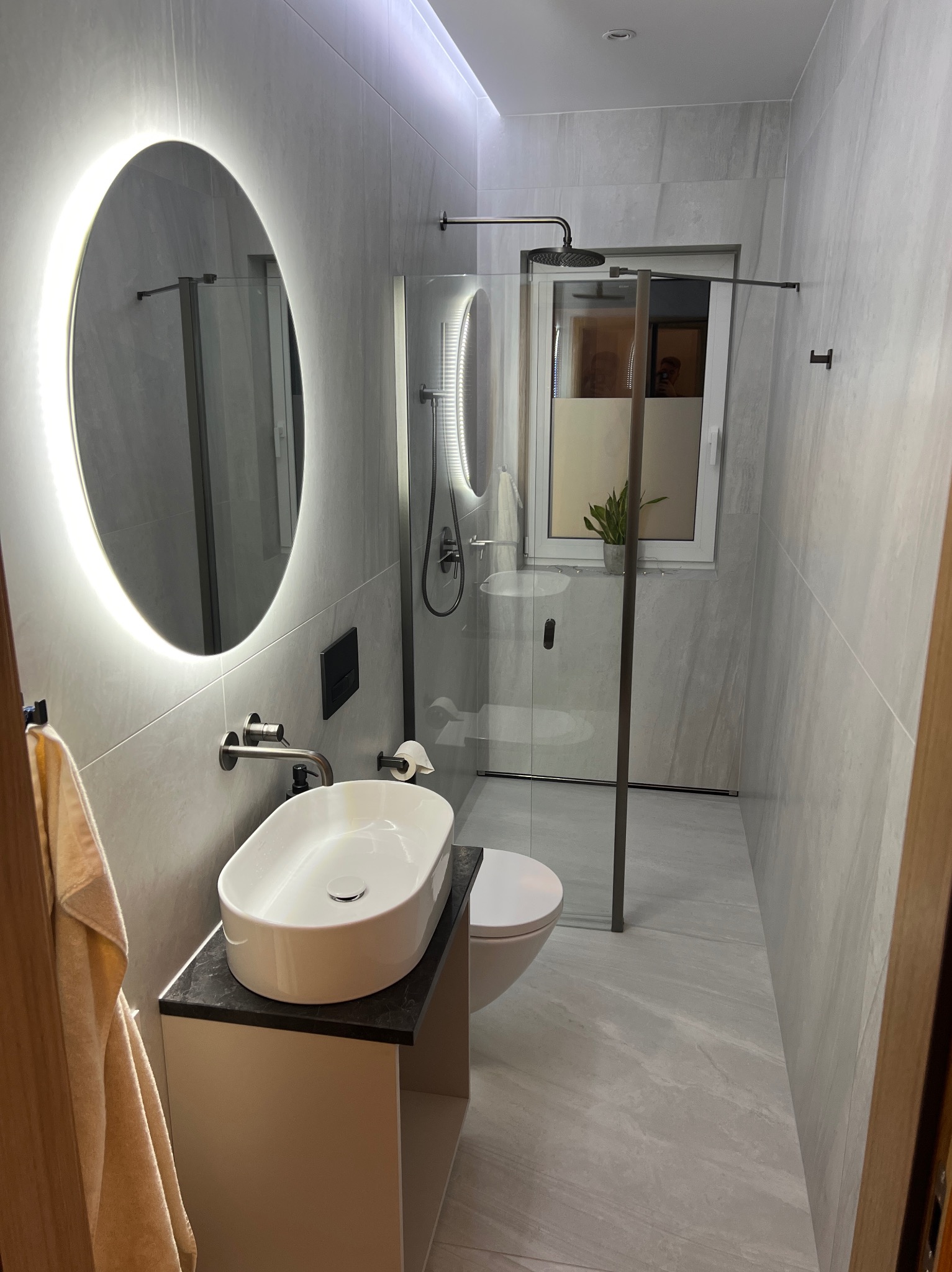 Nowoczesna łazienka z owalnym lustrem LED, kabiną prysznicową typu walk-in i umywalką nablatową na szafce. Szare płytki imitujące kamień na ścianach i podłodze.