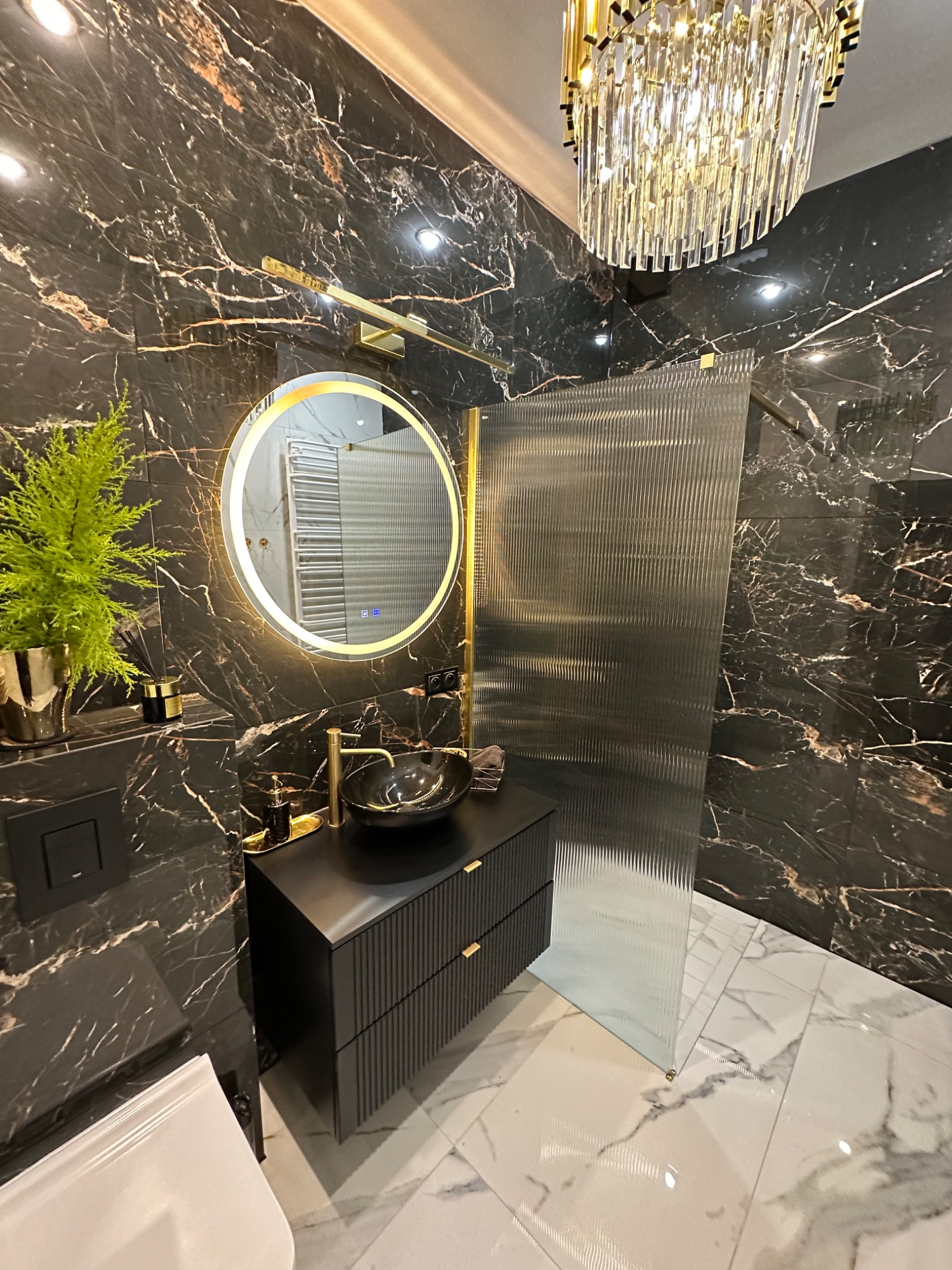 Ekskluzywna łazienka w czerni i złocie: marmurowe ściany, okrągłe lustro z podświetleniem LED, czarna umywalka nablatowa i szklana kabina prysznicowa.