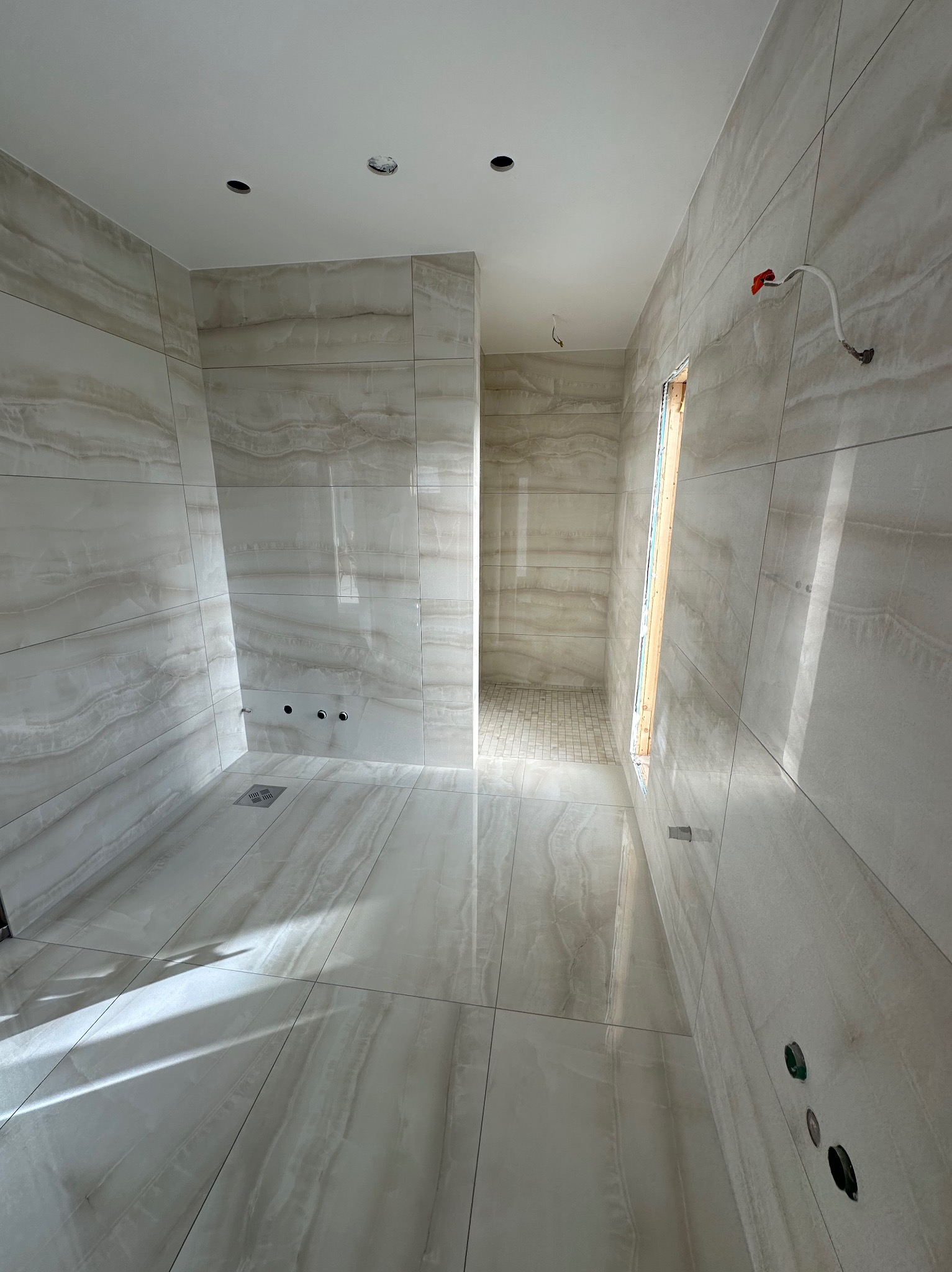 Wykończona łazienka w jasnych płytkach imitujących marmur. Widoczne podejścia wodne, odpływ liniowy i otwór okienny. Minimalistyczny design.