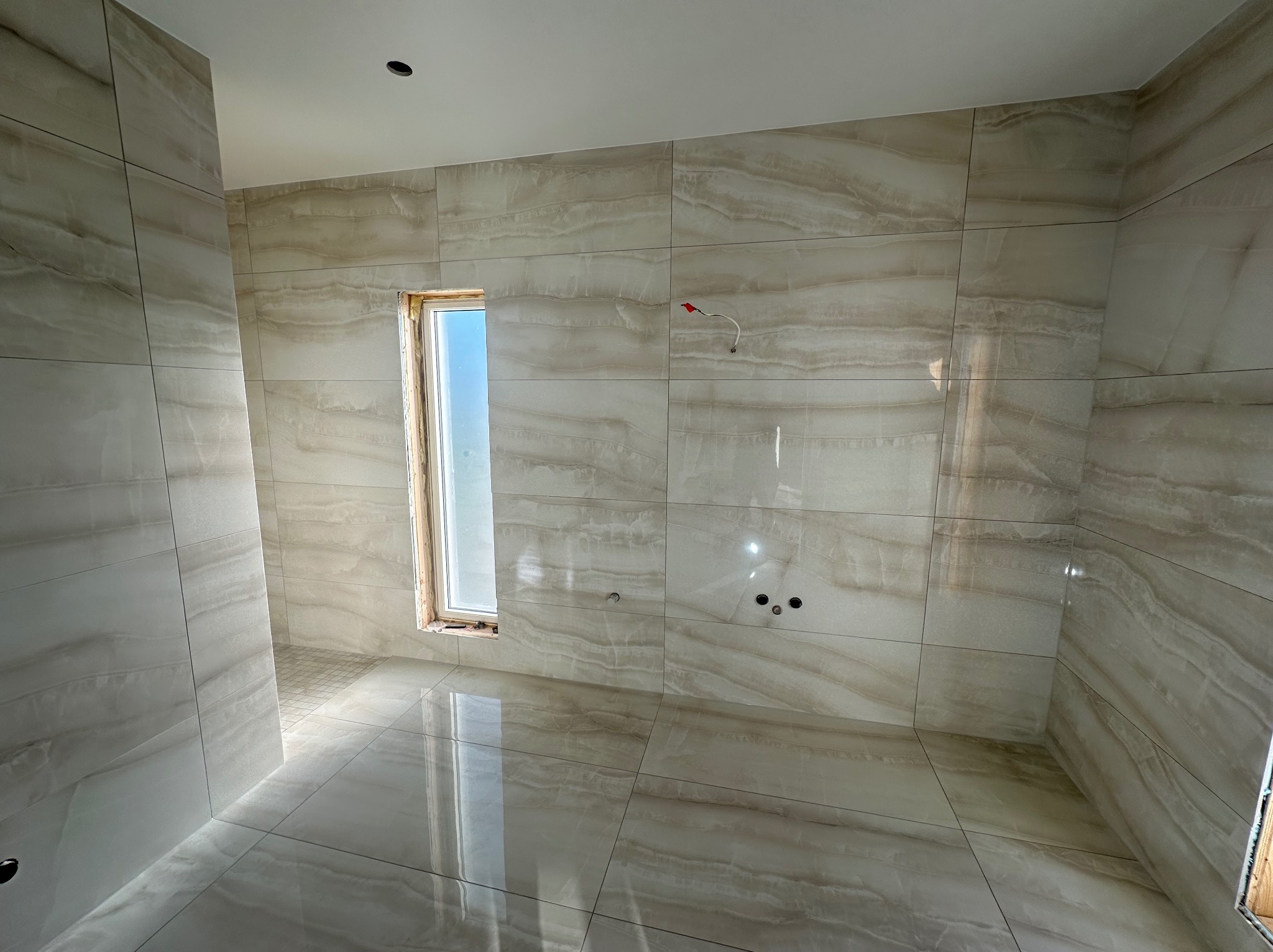 Jasna, wykafelkowana łazienka z oknem w drewnianej ramie. Widoczne otwory na armaturę i wystające przewody. Połyskująca podłoga odbija światło.