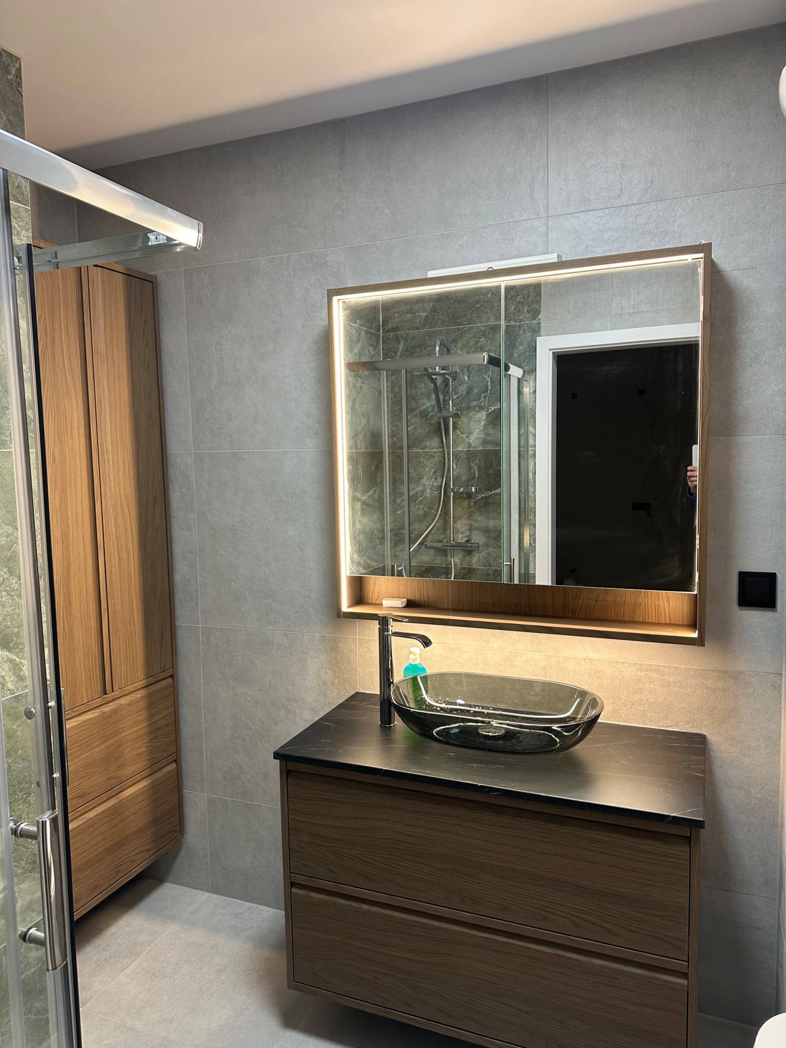 Nowoczesna łazienka z szarą glazurą, drewnianą szafką pod umywalkę z czarnym blatem i podświetlanym lustrem. Kabina prysznicowa z elementami marmuru.