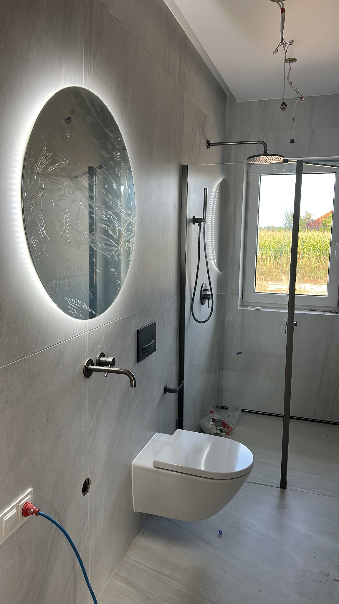 Nowoczesna łazienka w szarościach z podświetlanym lustrem, wiszącą toaletą i kabiną prysznicową typu walk-in. Widoczne niedokończone prace elektryczne.