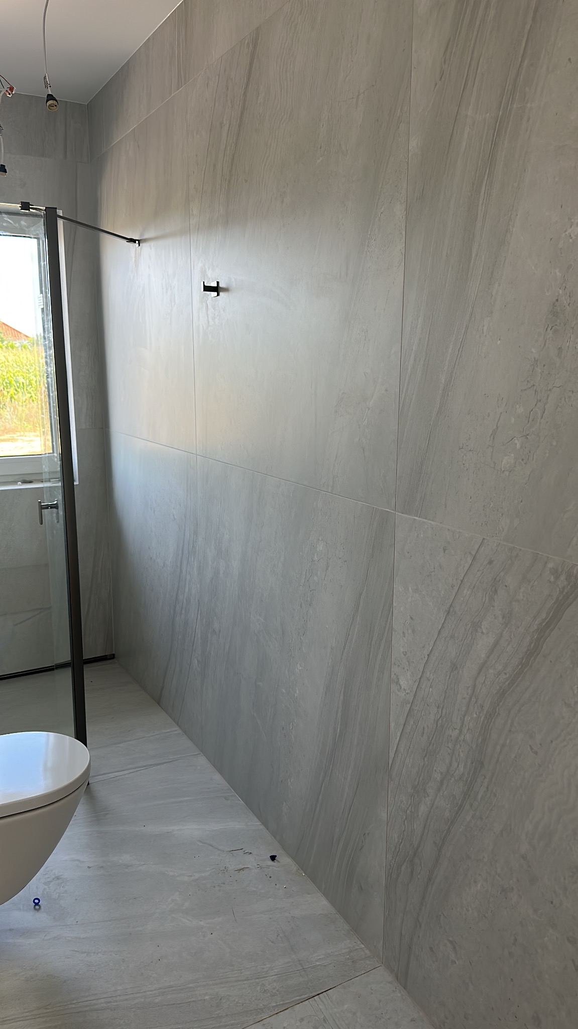 Wykończona łazienka z szarymi płytkami imitującymi kamień na ścianach i podłodze. Kabina prysznicowa z czarnymi elementami. Widoczna toaleta.