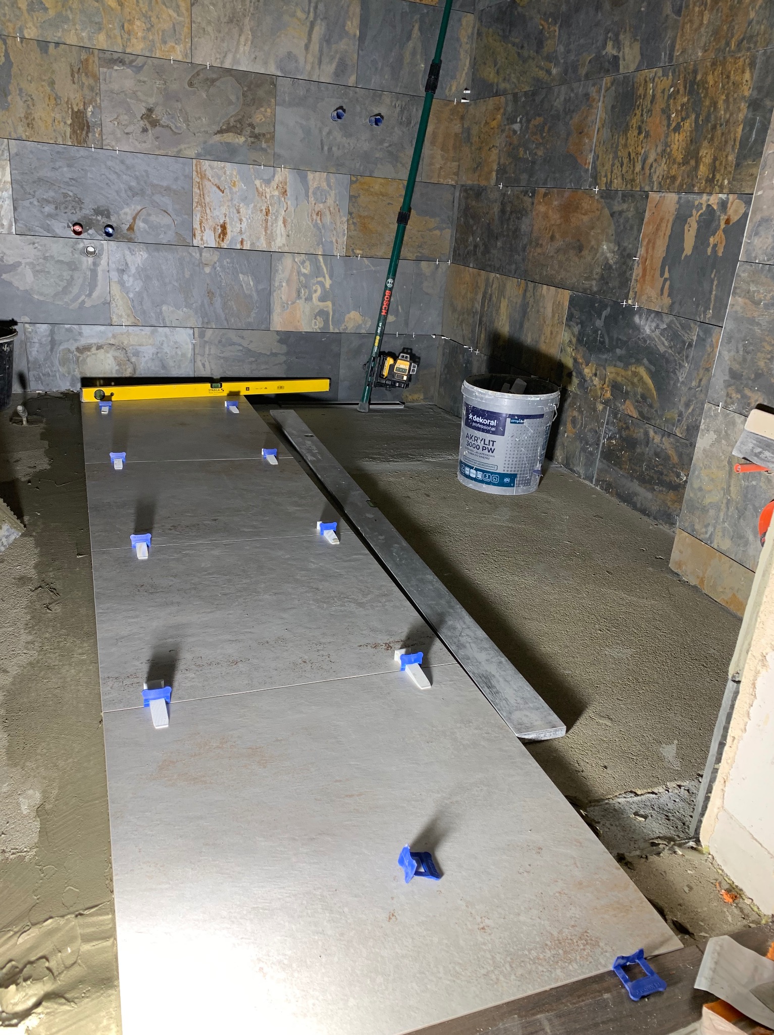 Układanie płytek w łazience z użyciem poziomicy i systemu poziomowania. Widoczny kubeł z zaprawą i laserowy niwelator. Ściany wyłożone płytkami imitującymi kamień.