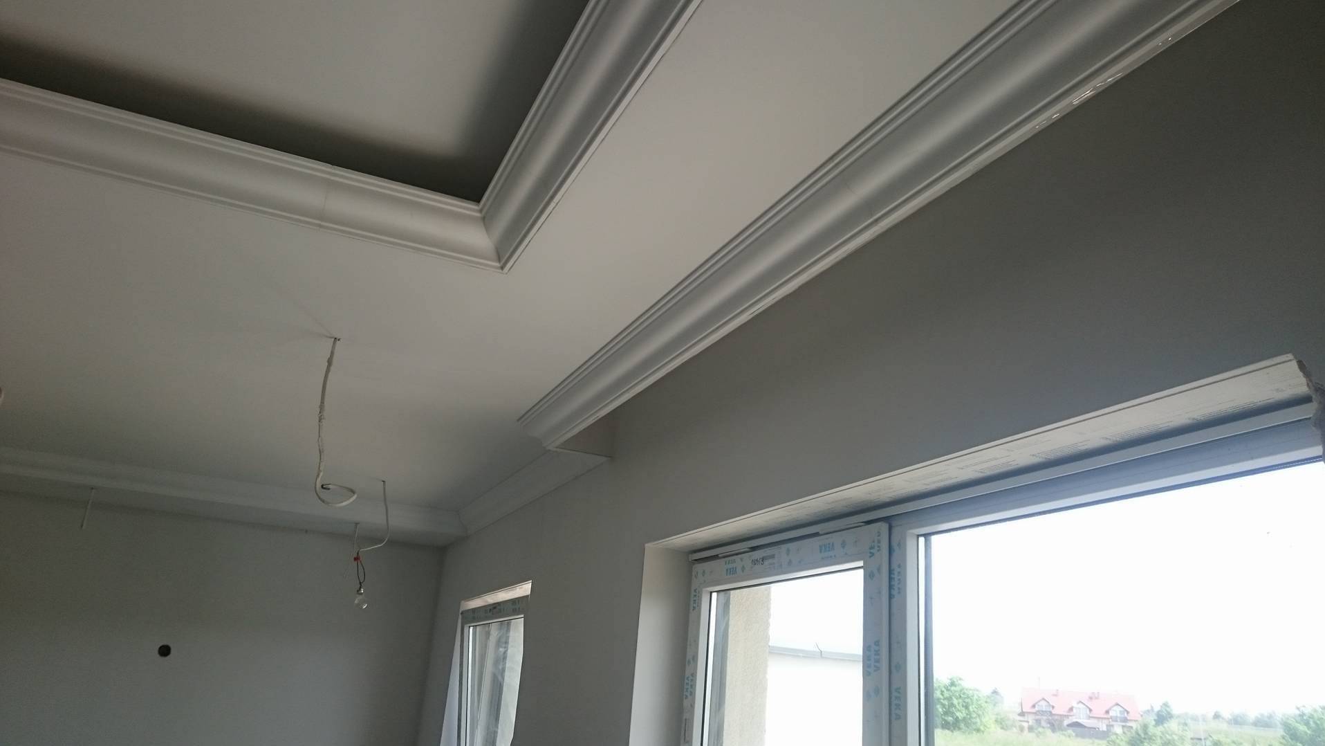 Wykończone wnętrze: biały sufit z dekoracyjnymi listwami, szare ściany i okno PCV z widokiem na zewnątrz. Wystające kable elektryczne.