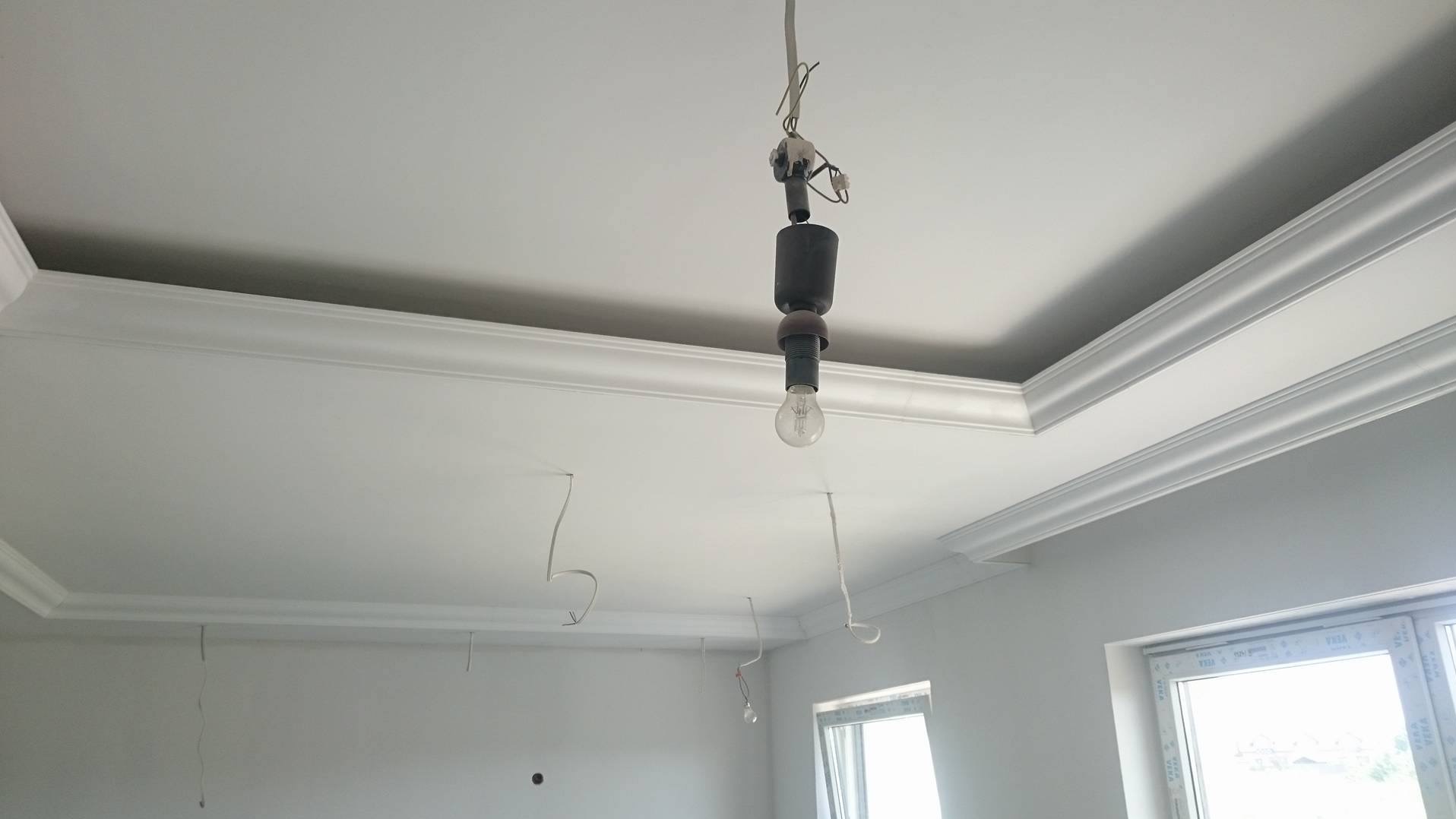 Biały sufit z dekoracyjną sztukaterią i wiszącą żarówką. Widoczne fragmenty ścian i okna. Wnętrze w trakcie wykańczania.
