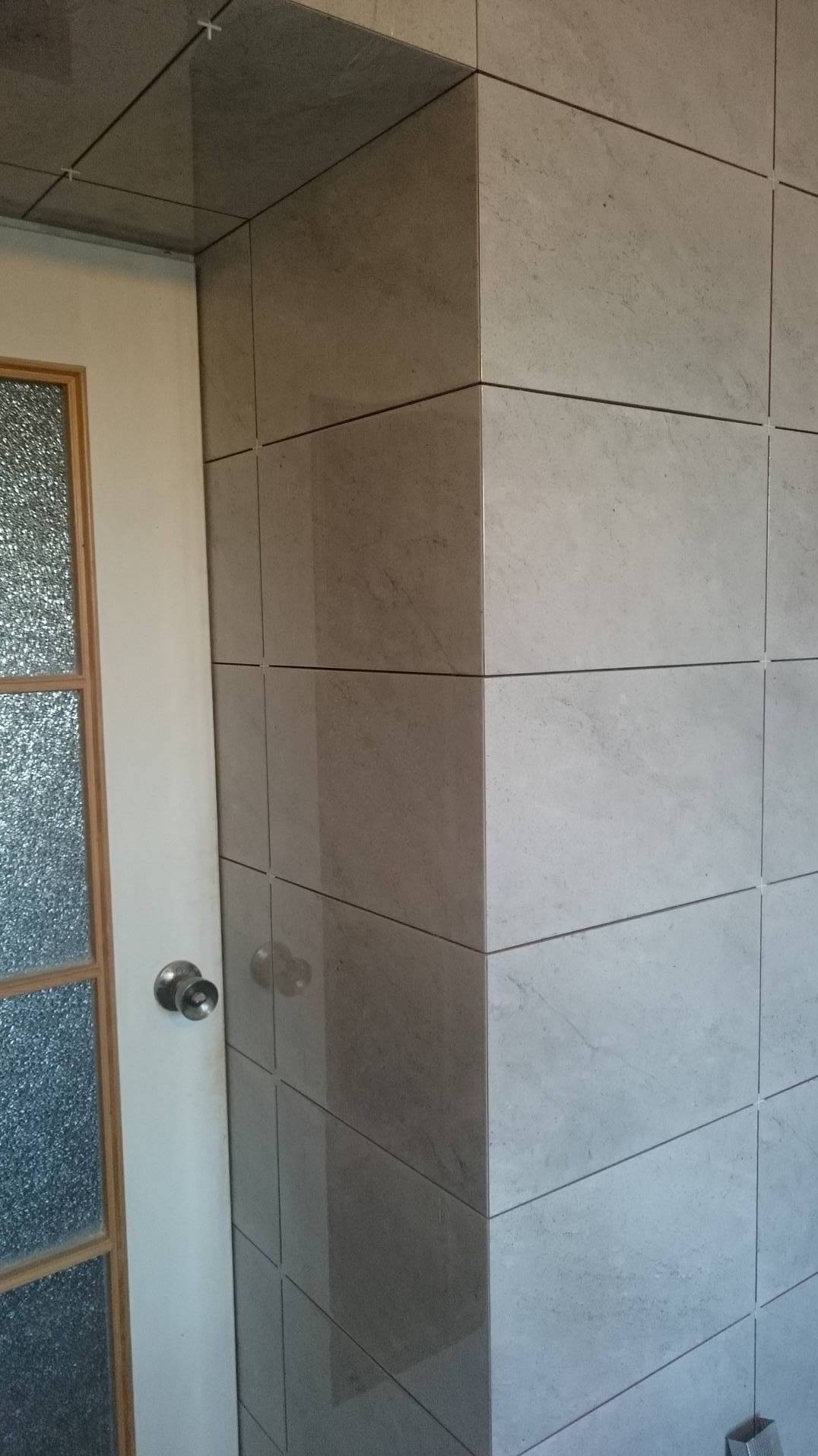 Wykończony narożnik łazienki z szarymi, matowymi płytkami. Widoczna biała ościeżnica drzwi z szybą ornamentową i chromowaną klamką. Estetyczne połączenia płytek.