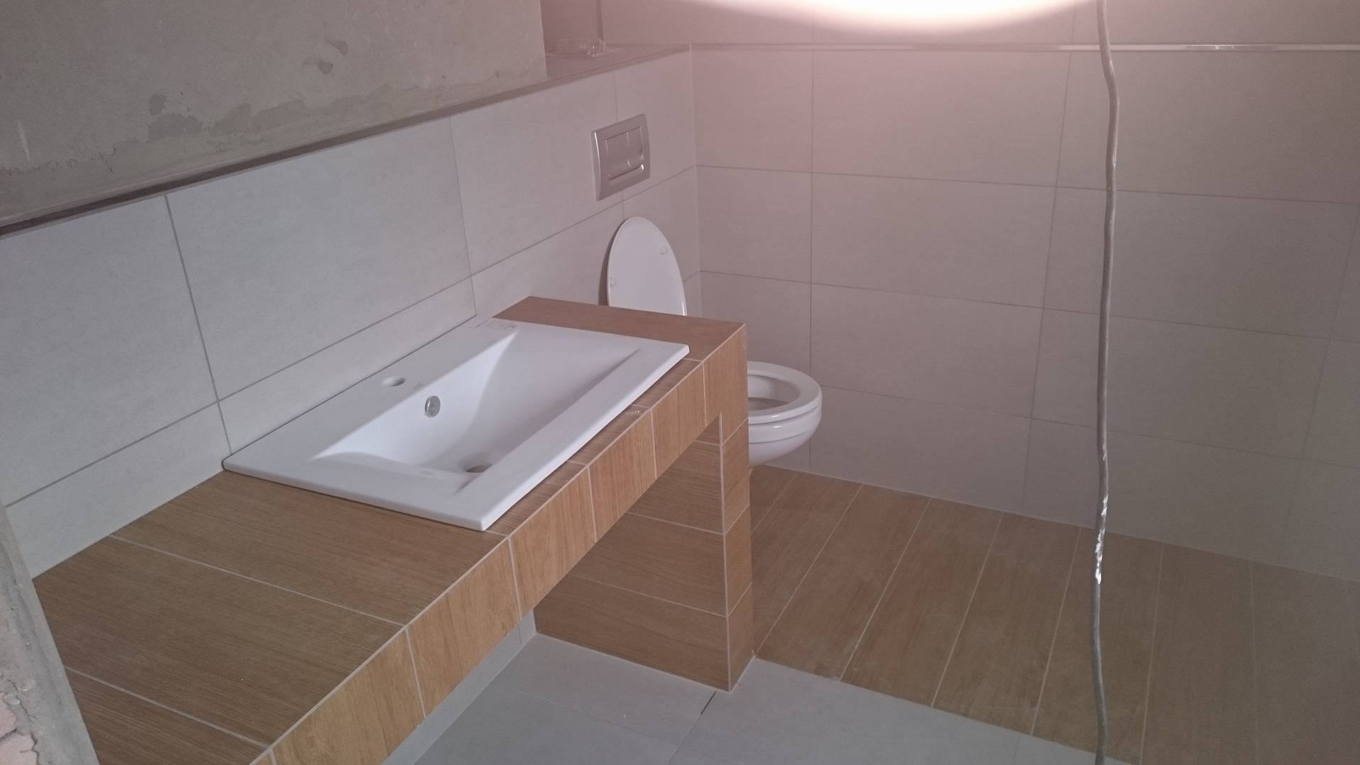 Wykończona łazienka z umywalką wpuszczoną w blat z płytek imitujących drewno, toaleta w tle, jasne płytki na ścianach i podłodze, surowy fragment ściany.