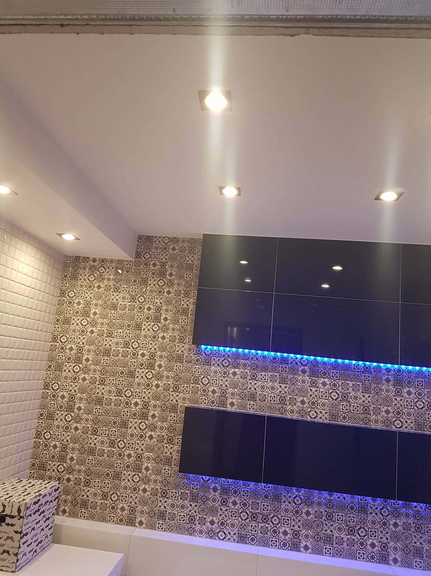 Wykończona ściana z mozaiką i czarnymi szafkami podświetlonymi niebieskim LED, oświetlenie sufitowe punktowe, białe płytki na ścianie, dekoracyjny kosz.