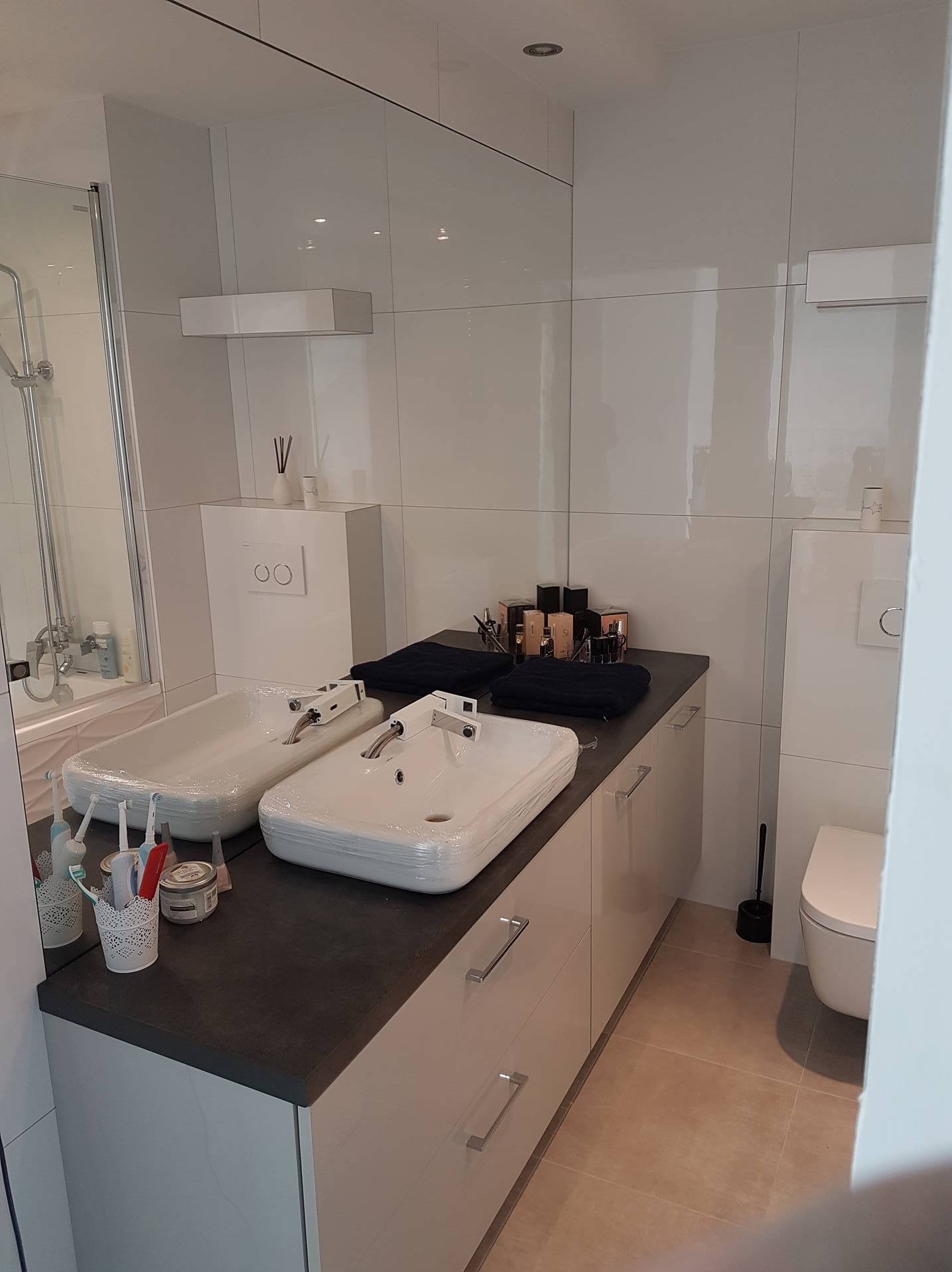 Nowoczesna łazienka z dwoma białymi umywalkami na ciemnym blacie, duże lustro, białe płytki i szafki. Widoczna toaleta i akcesoria łazienkowe.