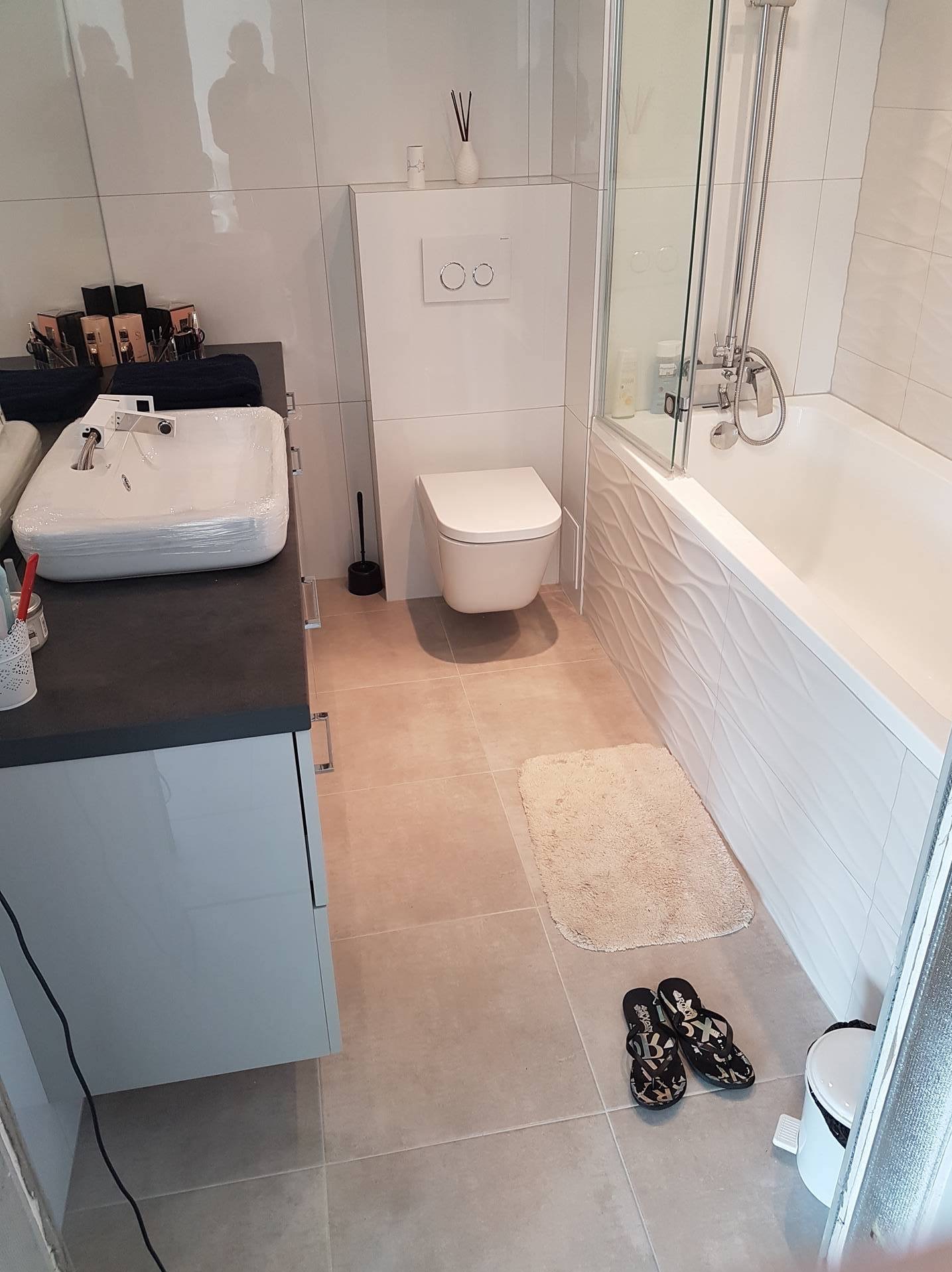 Jasna łazienka po remoncie z umywalką owiniętą folią ochronną, beżowe płytki podłogowe i biała wanna z parawanem.