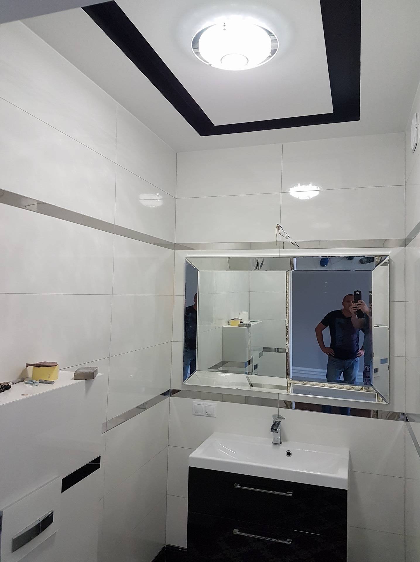 Biała łazienka z czarnymi akcentami: kafelki, sufit. Lustro z fazowanymi krawędziami odbija osobę. Wisząca szafka z umywalką w kolorze czarnym.