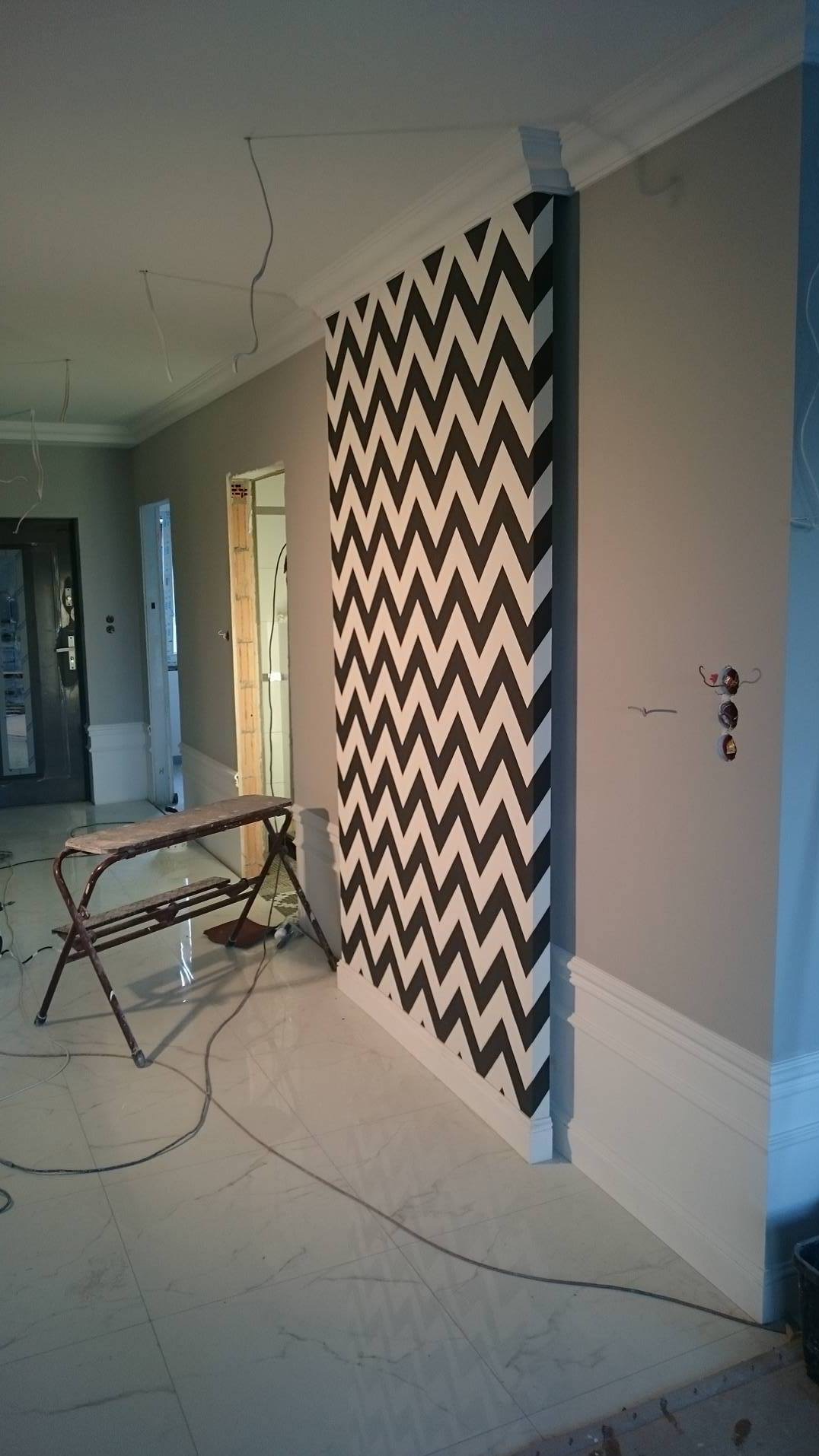 Ściana z wzorem w zygzaki w wykończonym wnętrzu, widoczne listwy przypodłogowe, drabina malarska i kable. W tle widoczne drzwi i fragment drugiego pomieszczenia.