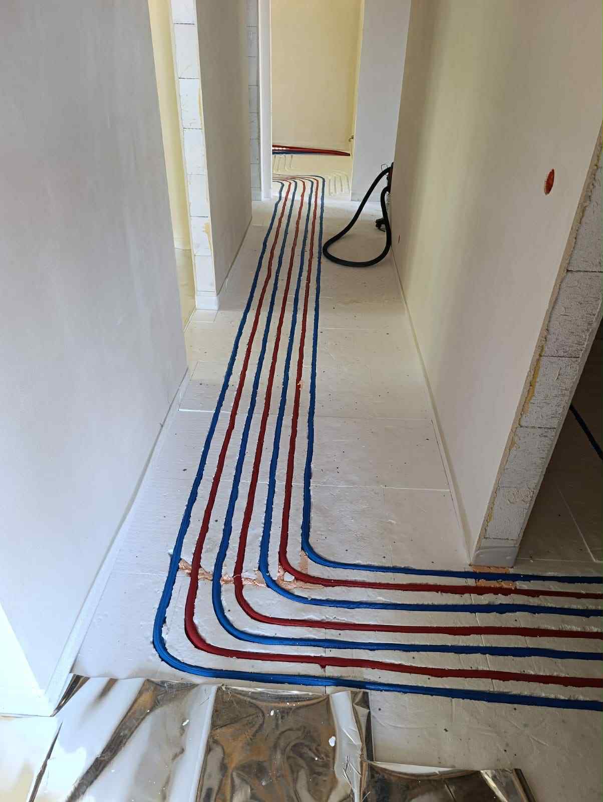 Biała podłoga z izolacją i ułożonymi rurami ogrzewania podłogowego w kolorze niebieskim i czerwonym, widok z góry w korytarzu w trakcie prac instalacyjnych.