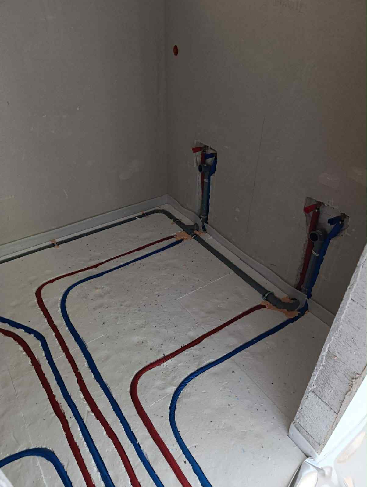 Instalacja ogrzewania podłogowego z rurami w kolorze niebieskim i czerwonym, ułożonymi na białej izolacji w surowym wnętrzu. Widoczne pionowe rury przy ścianie.