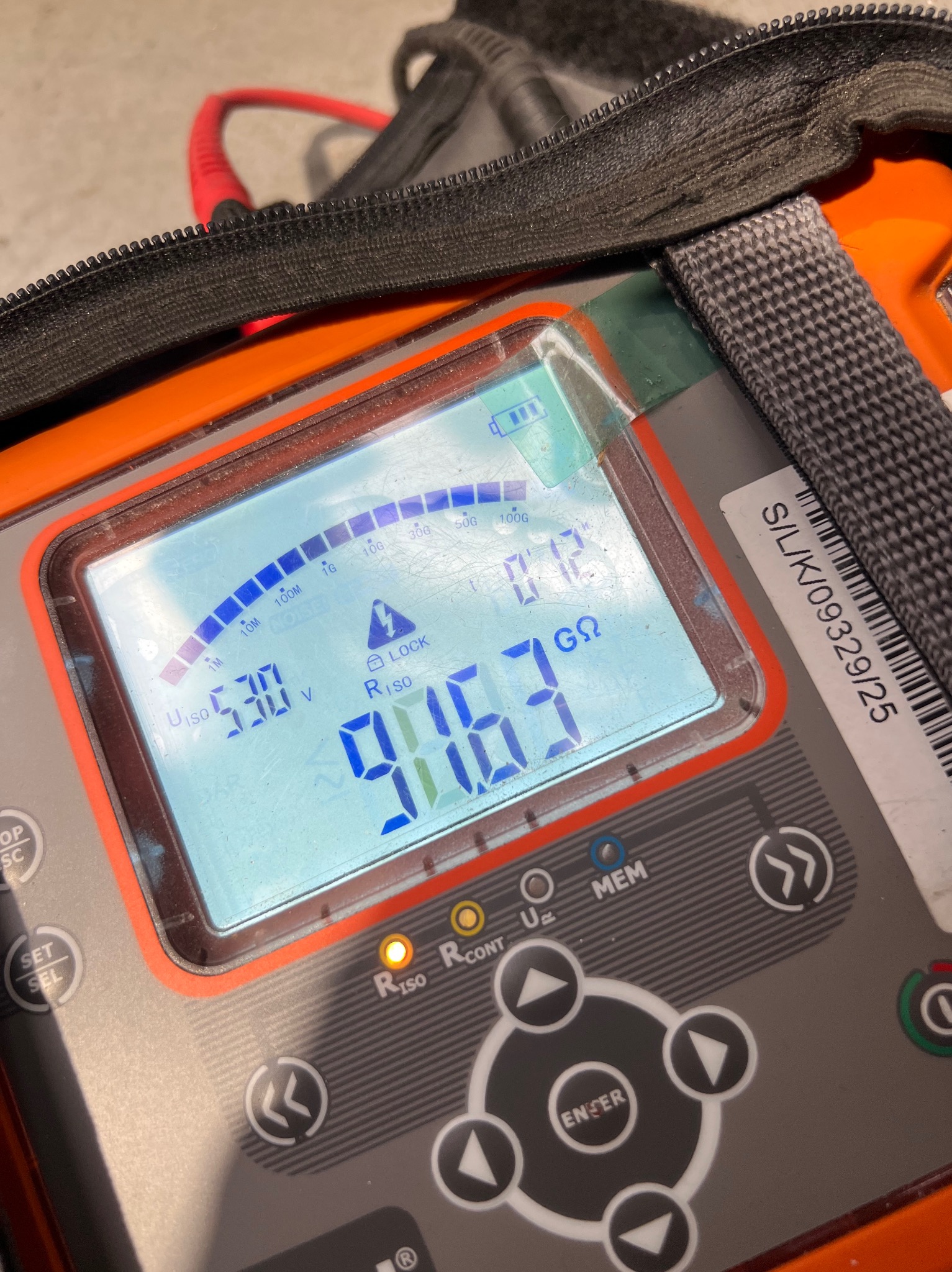 Miernik instalacji elektrycznych w pomarańczowej obudowie, wyświetlający wynik pomiaru rezystancji izolacji 916.3 GΩ, z widocznym fragmentem torby transportowej.