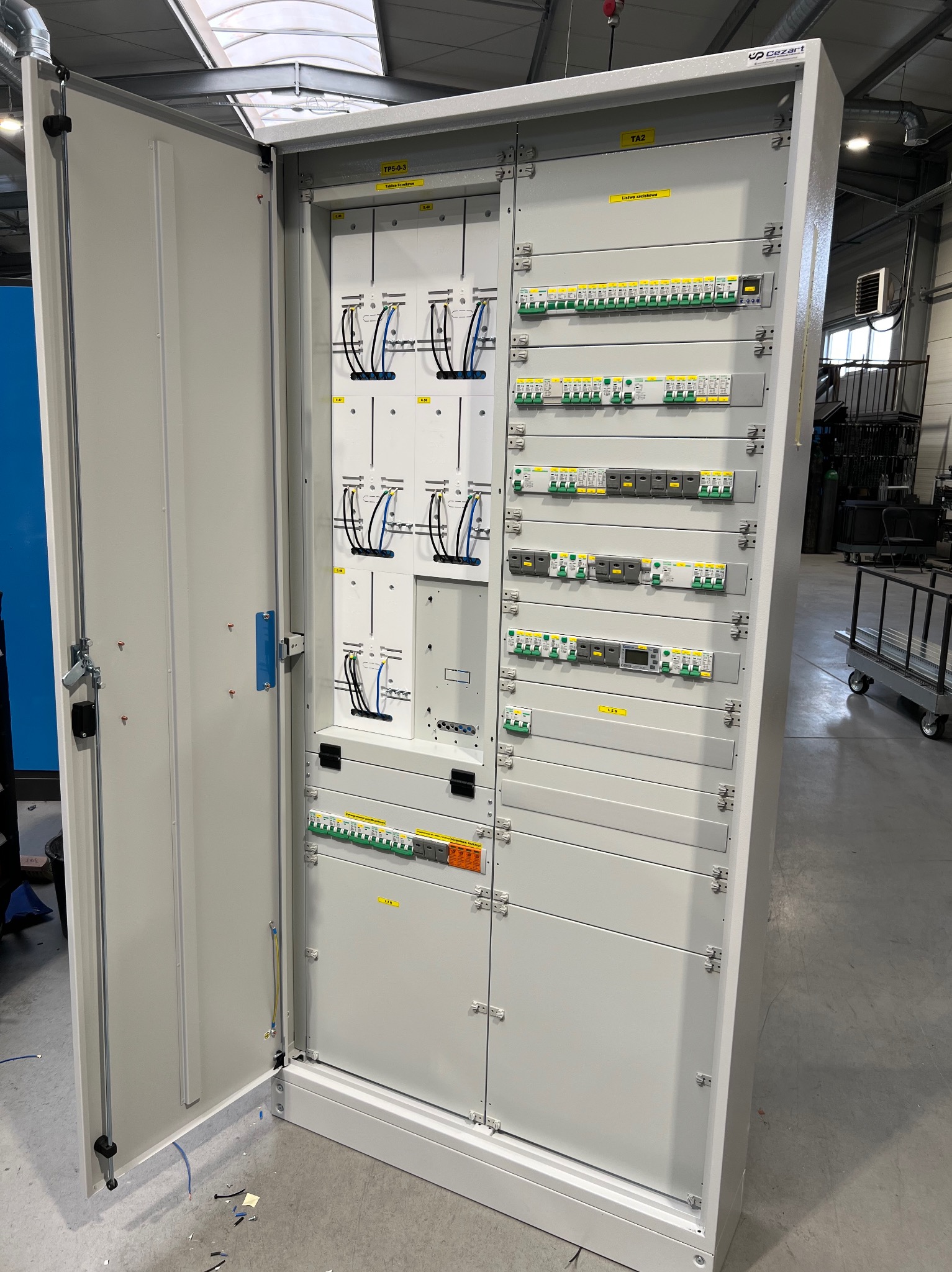 Otwarta szafa elektryczna z widocznymi bezpiecznikami, przewodami i elementami sterującymi, w przemysłowym otoczeniu. Widoczne żółte etykiety informacyjne.