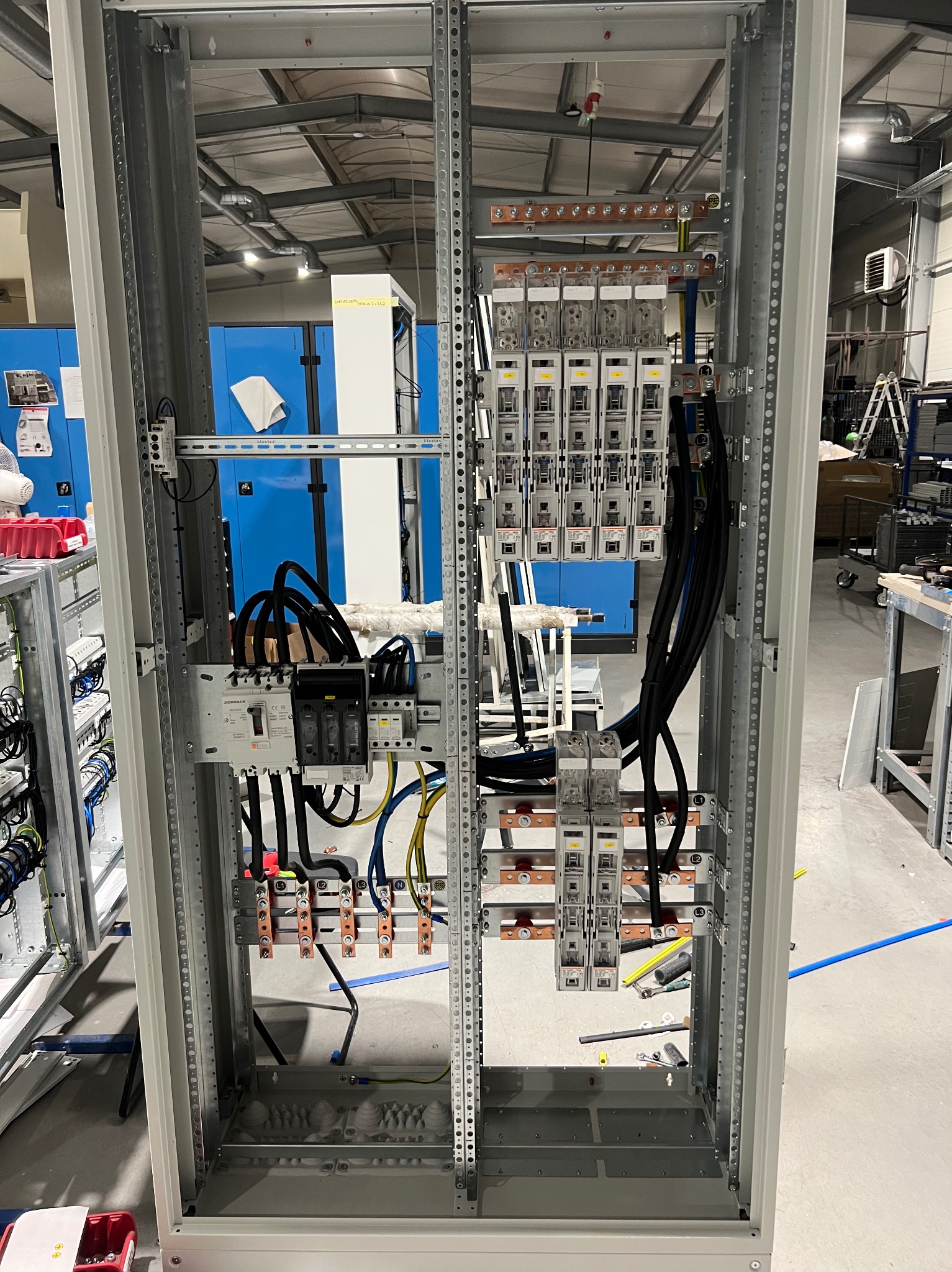 Rozdzielnica elektryczna w trakcie montażu: widoczne zabezpieczenia, okablowanie, szyny miedziane. Metalowa konstrukcja w jasnej hali produkcyjnej.