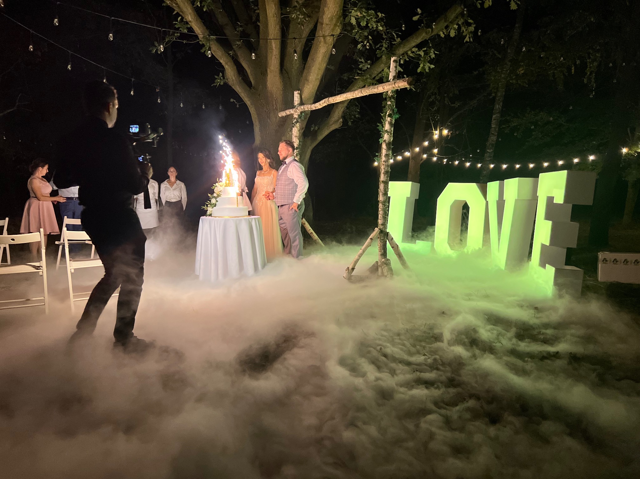 Para młoda kroi tort z fontannami iskier podczas wesela plenerowego z dekoracjami LOVE i efektem ciężkiego dymu, filmowana przez kamerzystę.