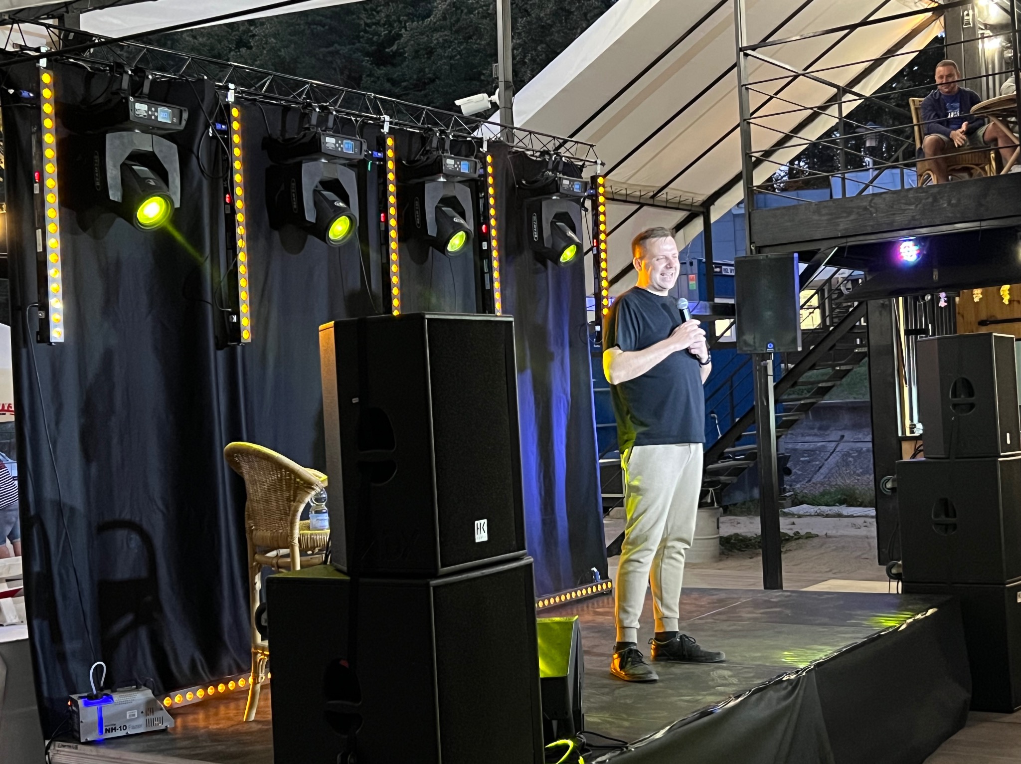 Uśmiechnięty konferansjer na scenie z nagłośnieniem i oświetleniem. W tle konstrukcja stalowa z osobą siedzącą na górze. W tle ciemne niebo.