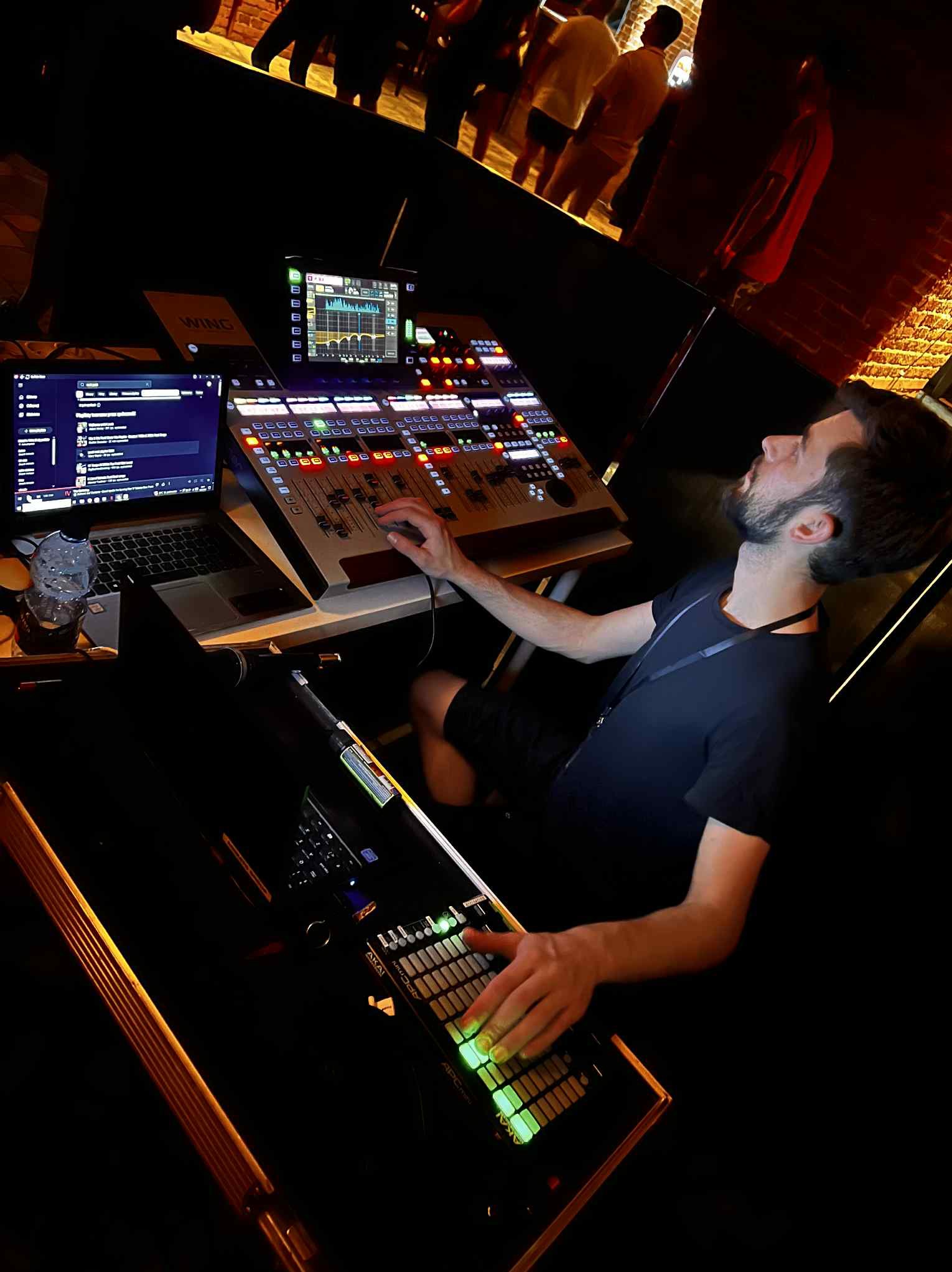 Mężczyzna obsługuje konsoletę mikserską Wing i kontroler MIDI Akai APC mini w ciemnym otoczeniu, prawdopodobnie podczas wydarzenia z widocznymi sylwetkami osób w tle.